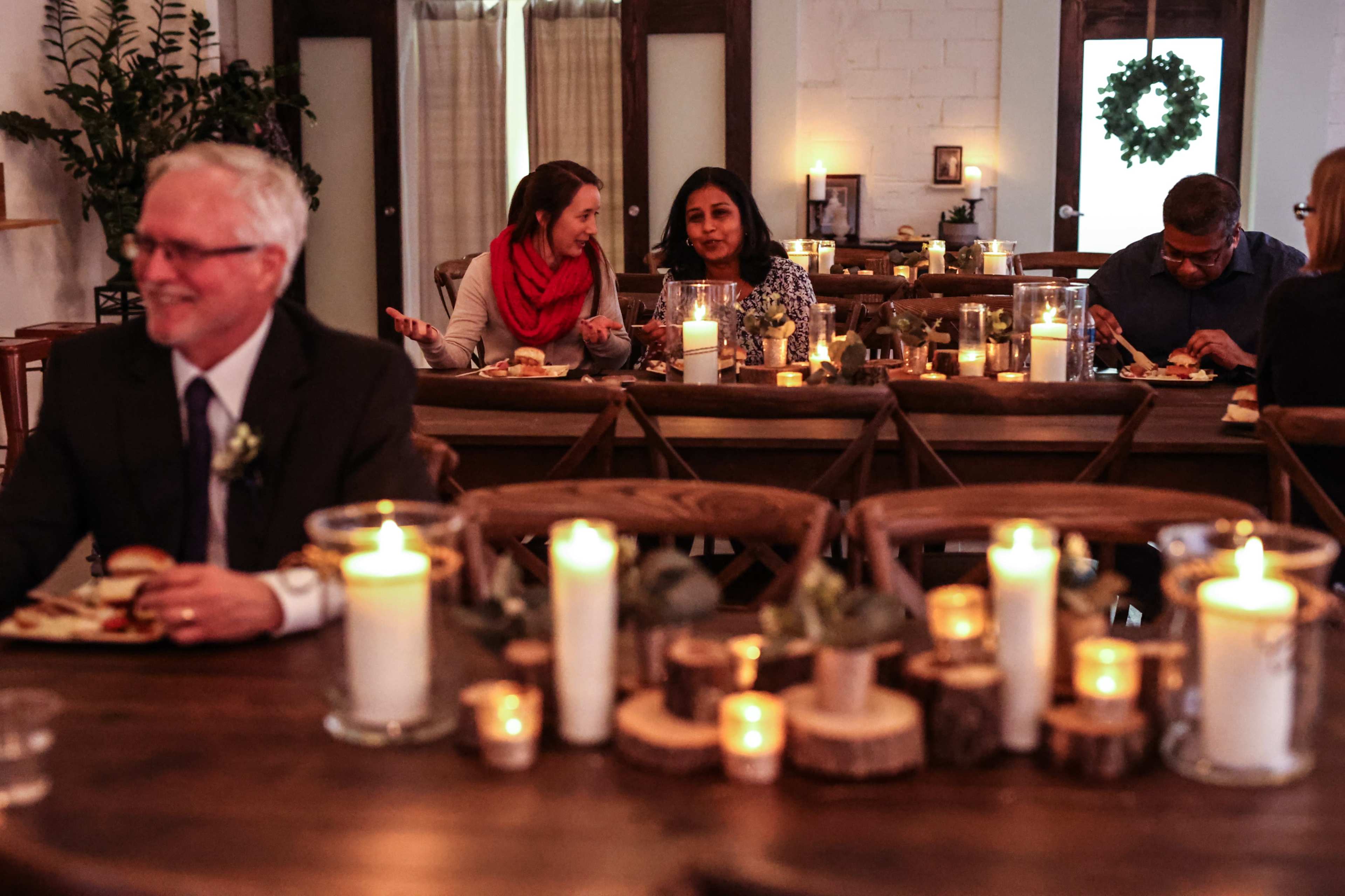 A cozy dining room features guests seated at tables with candles, enjoying a meal, while a man smiles in the foreground.