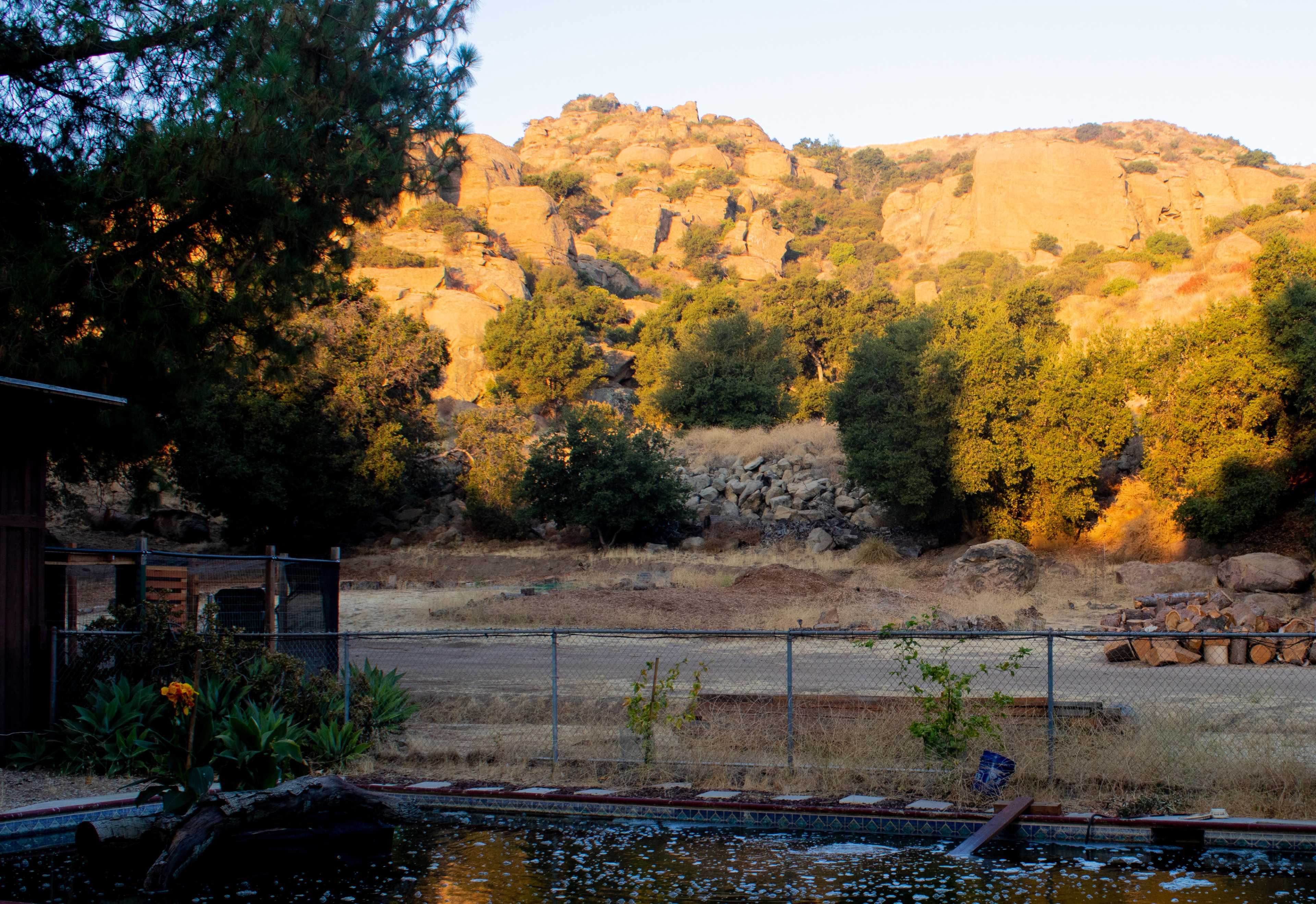 A dry, rocky landscape with a mountainous backdrop and scattered trees, bordered by a fenced area featuring a pool in the foreground.