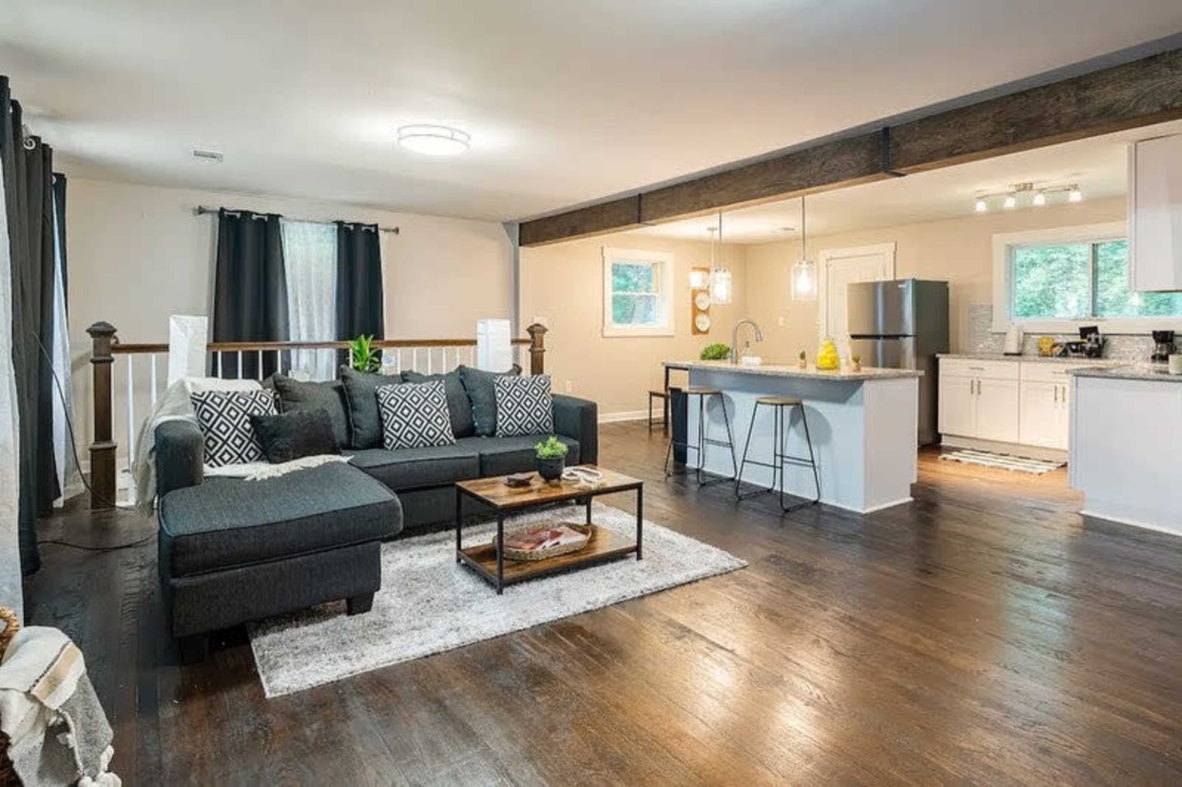A modern living room with a dark sectional sofa, a coffee table, and a kitchen area featuring white cabinets and a bar counter.