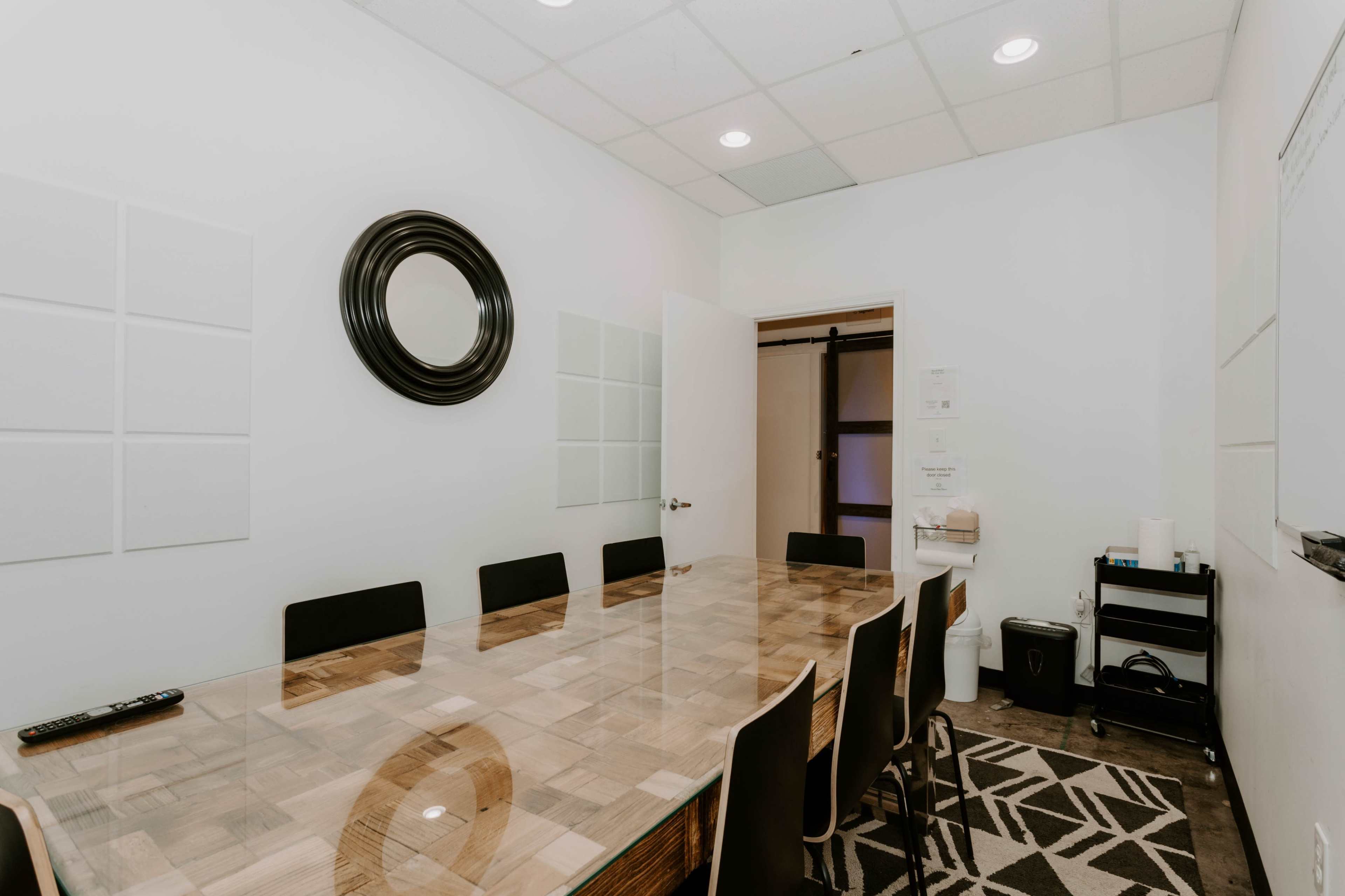 A conference room features a long wooden table surrounded by black chairs, with a mirror on the wall and a sliding door leading to an adjacent space.