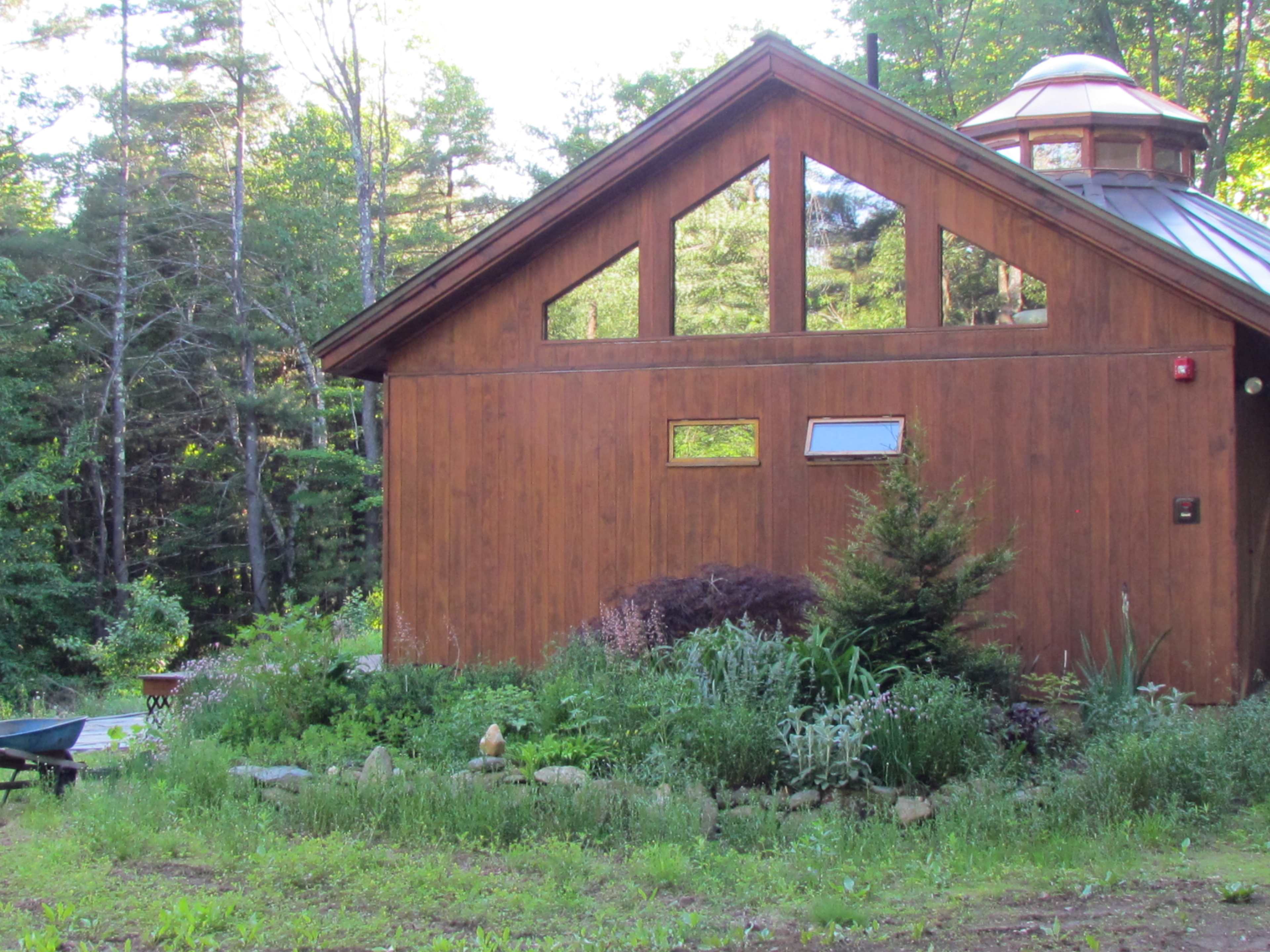 A wooden house with large windows is surrounded by a garden and trees in a wooded area.
