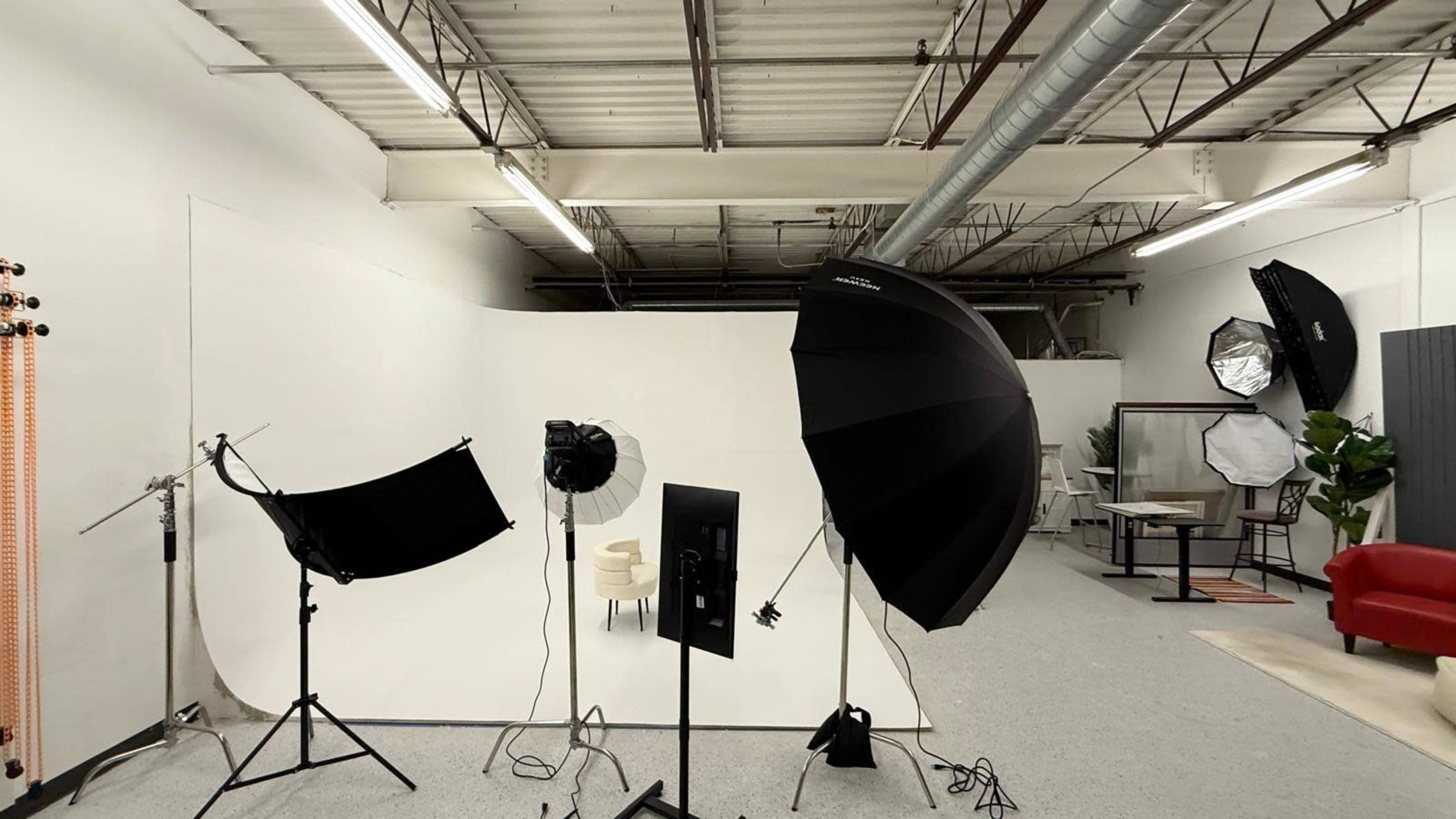 The image shows a photography studio with various lighting equipment and a white backdrop, alongside furniture and greenery in a spacious layout.
