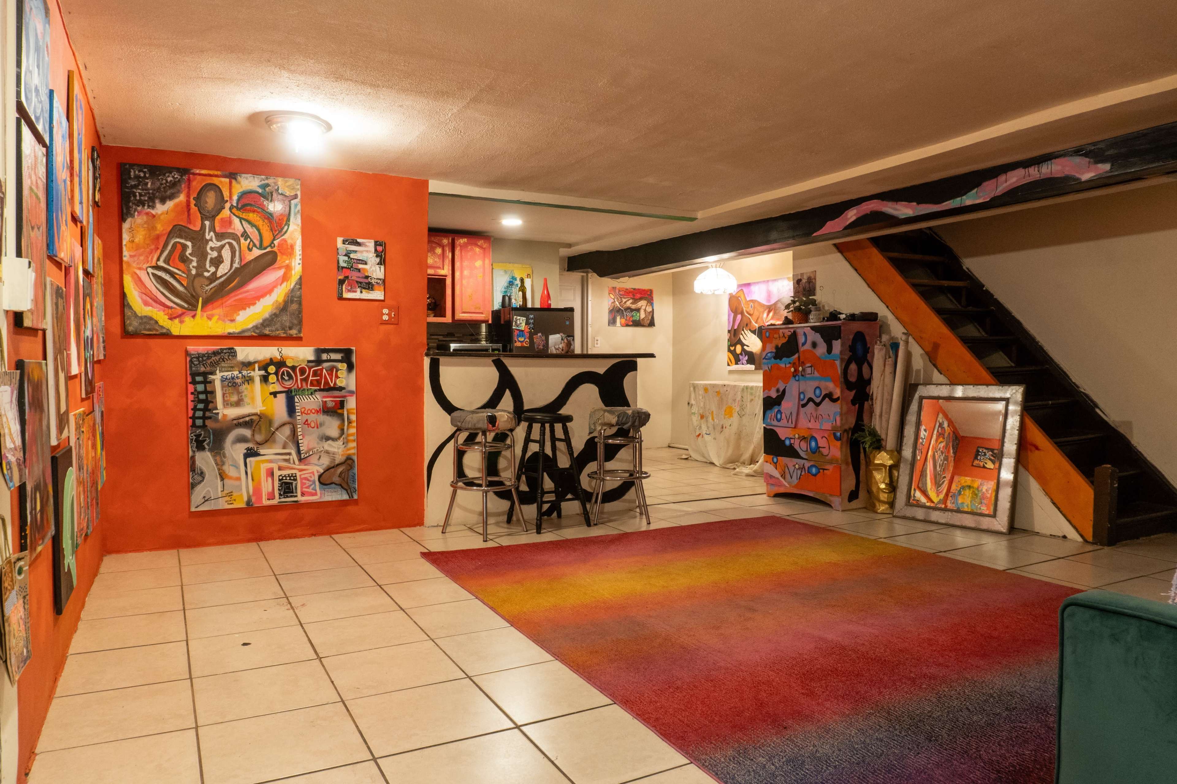 The image shows a colorful basement space with vibrant wall art, a small kitchenette in the background, and a staircase leading to an upper level.