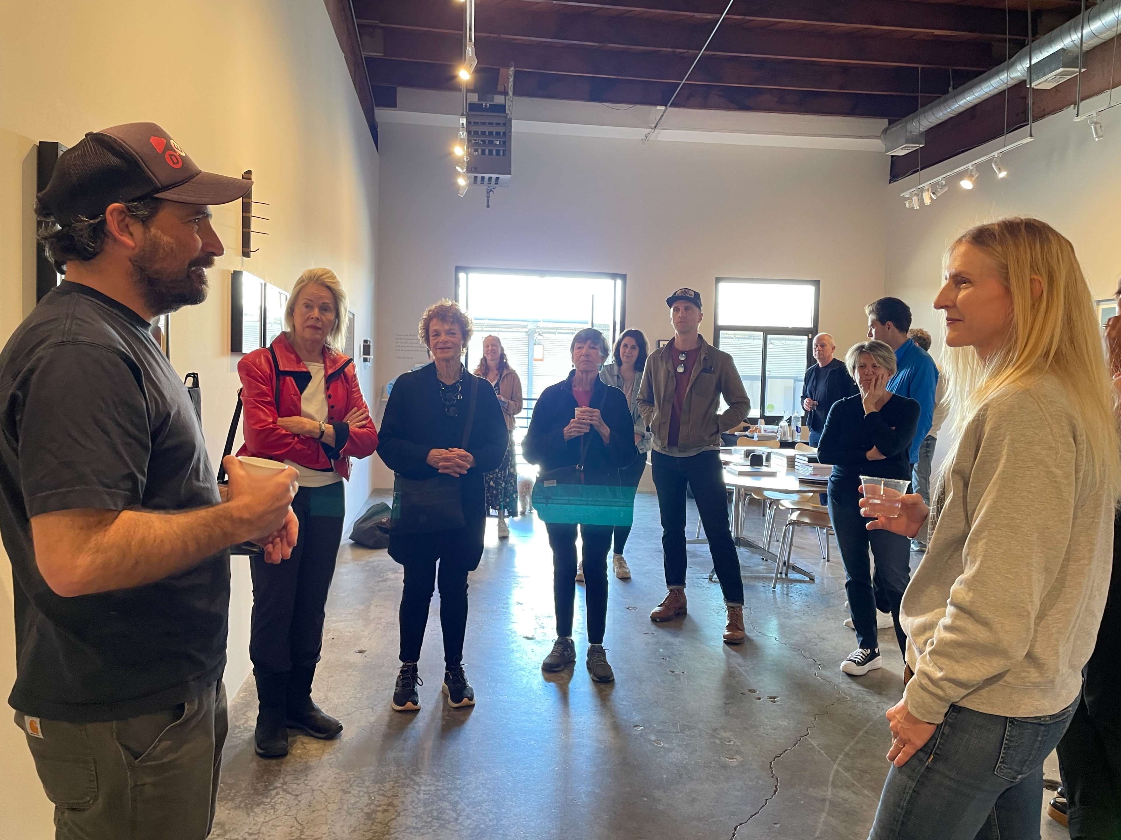 A group of people listens as a man speaks in a gallery setting with artwork on the walls.