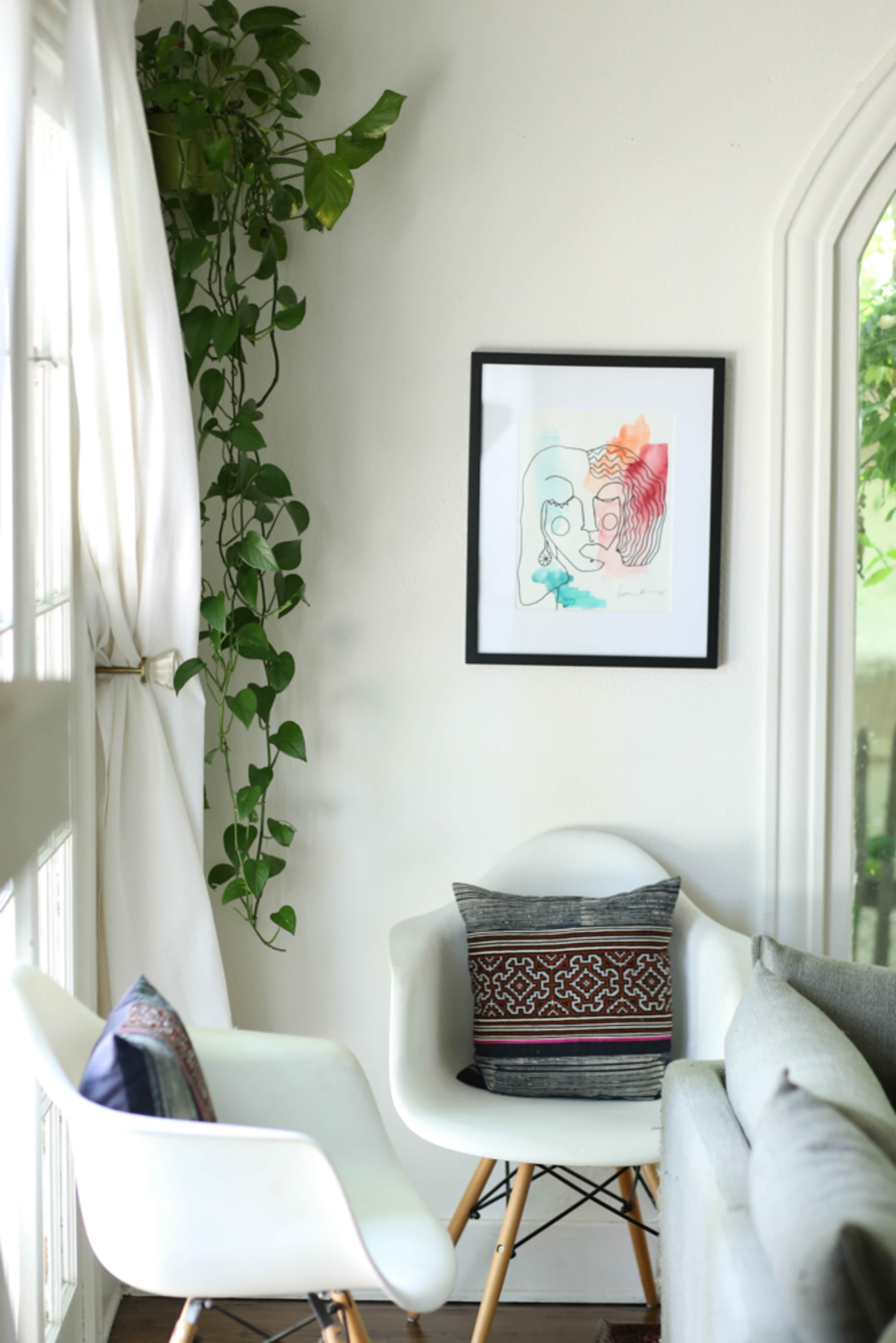A modern living space features a white chair with a patterned cushion, a gray sofa, and a framed artwork hanging on the wall, accompanied by a green plant on the side.