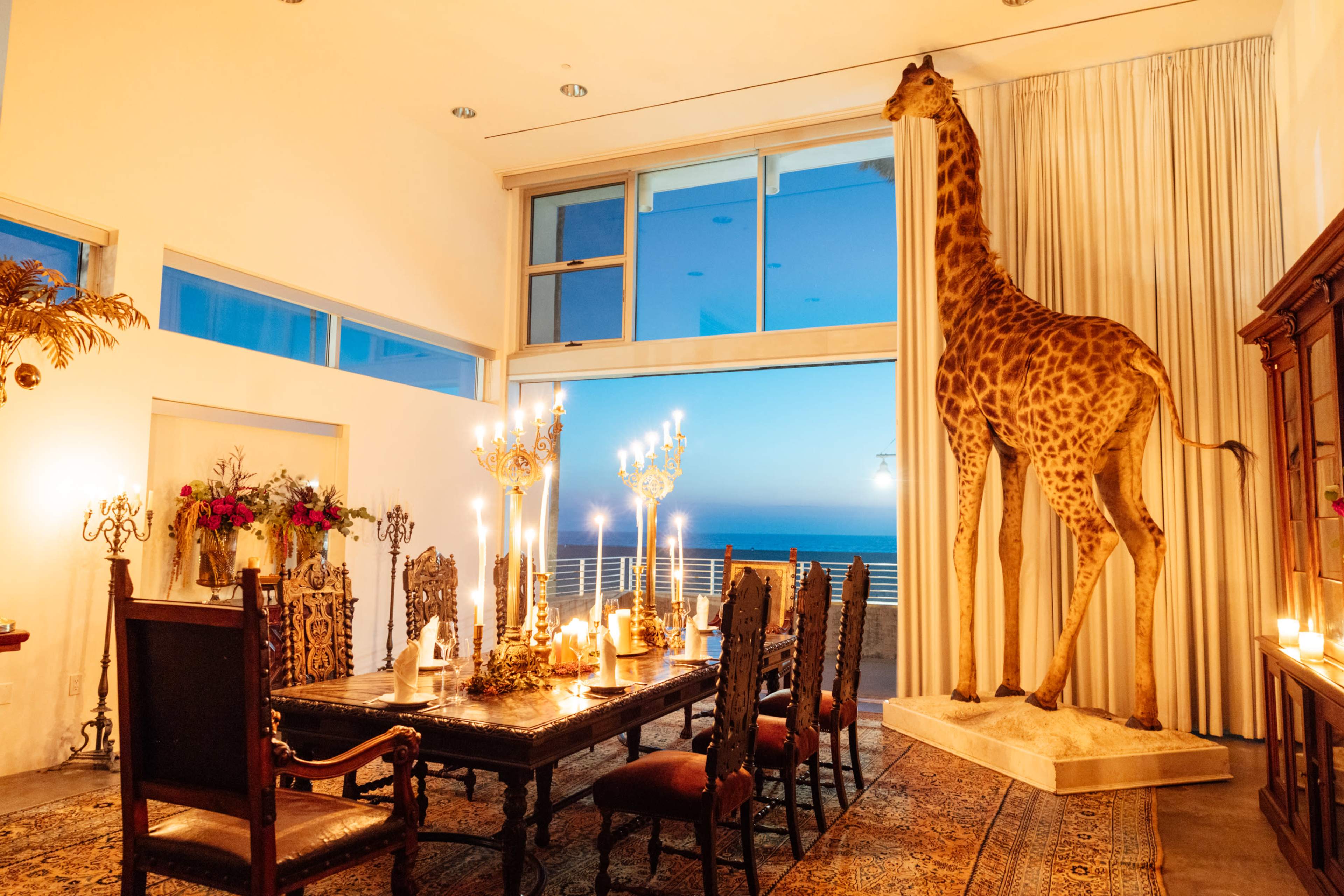 A lavish dining room features a large wooden table surrounded by chairs, with a mounted giraffe statue standing beside a window that showcases a view of the ocean at dusk.