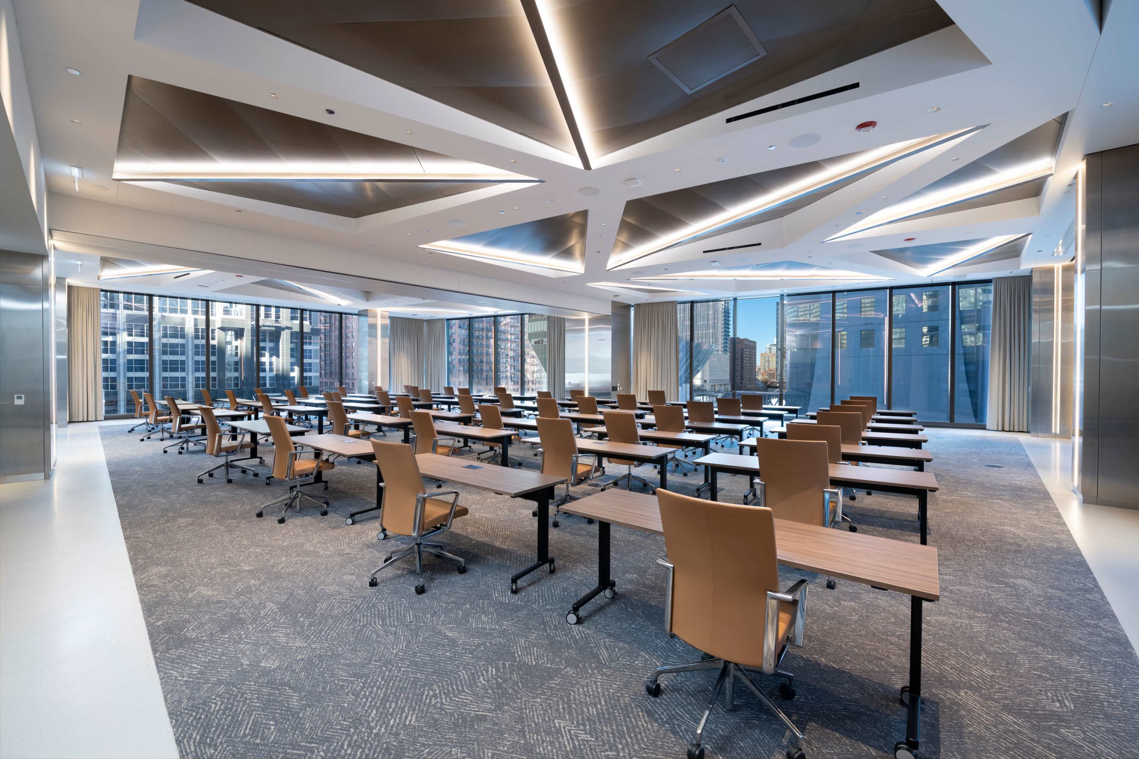 The image shows a modern conference room with rows of tables and chairs arranged in a spacious layout, featuring large windows that overlook a city skyline.
