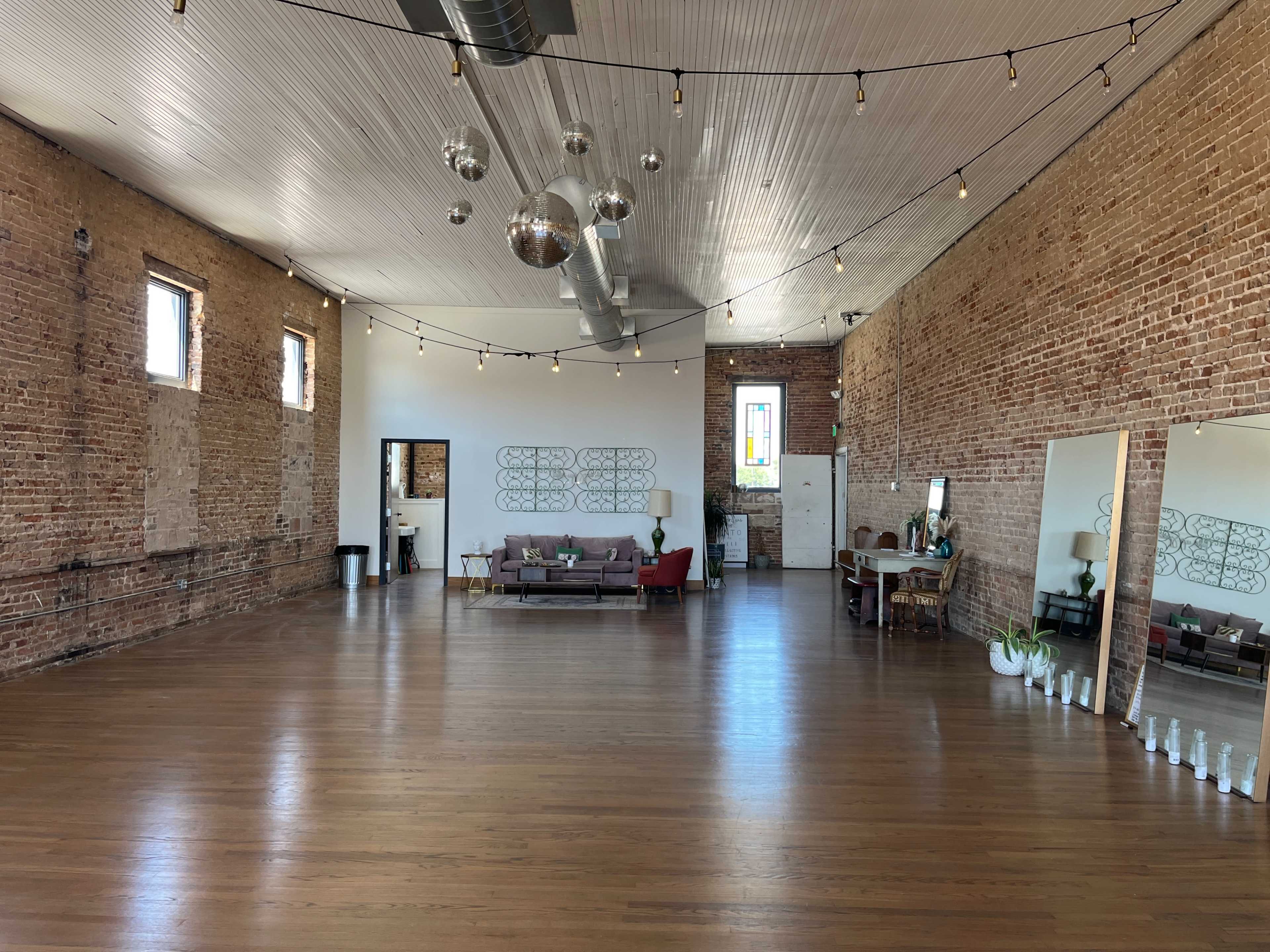 The spacious room features exposed brick walls, wooden flooring, and a mirrored section opposite a seating area with a couch and decor.