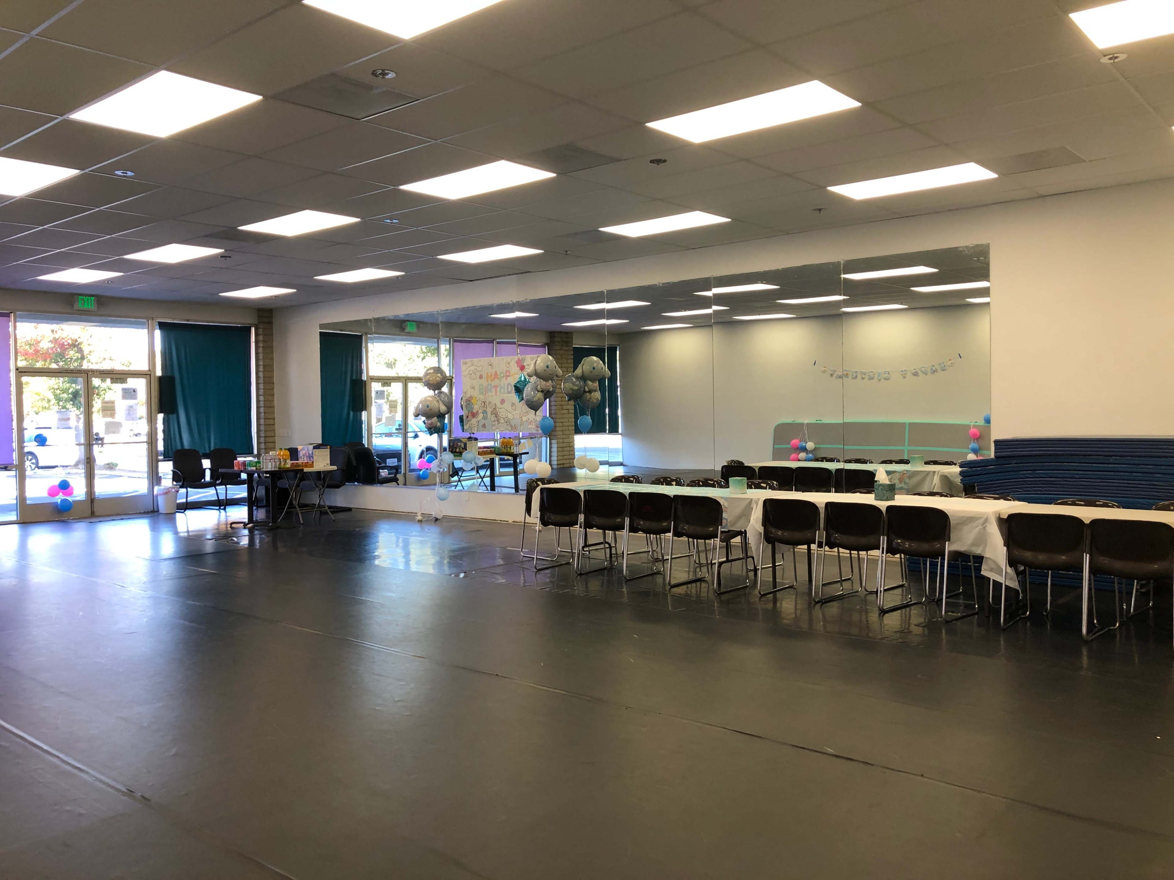 The image shows a spacious room with chairs arranged around a long table, large mirrors on one wall, and decorations for an event.