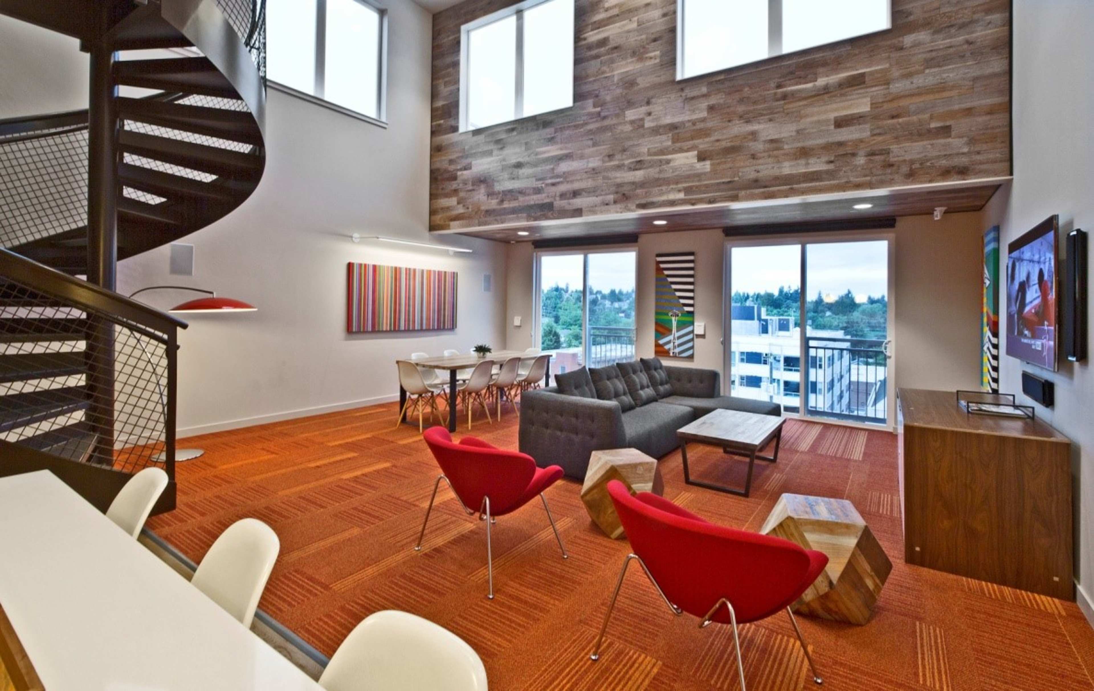 The image shows a modern living space with a spiral staircase, large windows, and a mix of seating areas, including a sectional sofa and red chairs.