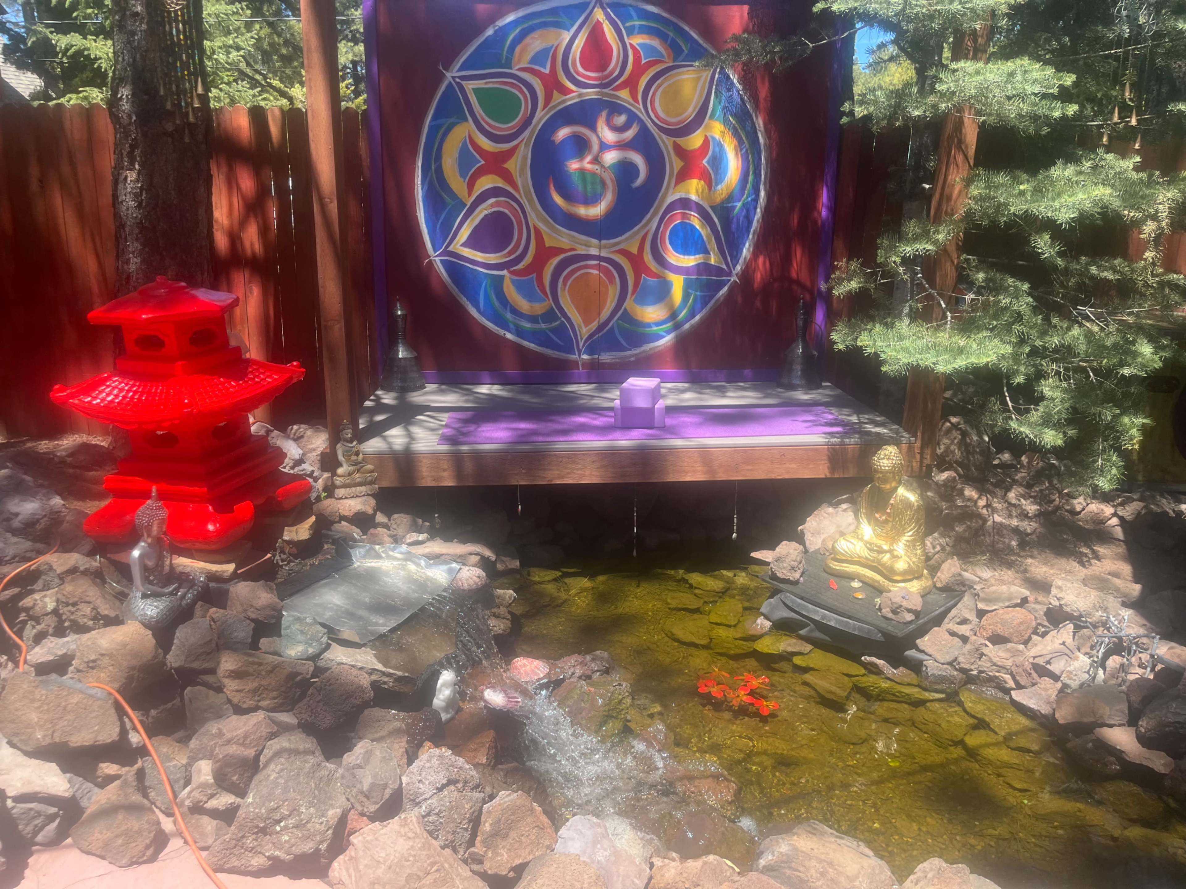 A tranquil garden scene featuring a pond with rocks, a gold statue of a seated Buddha, a purple backdrop with a circular symbol, and a red lantern beside the water.