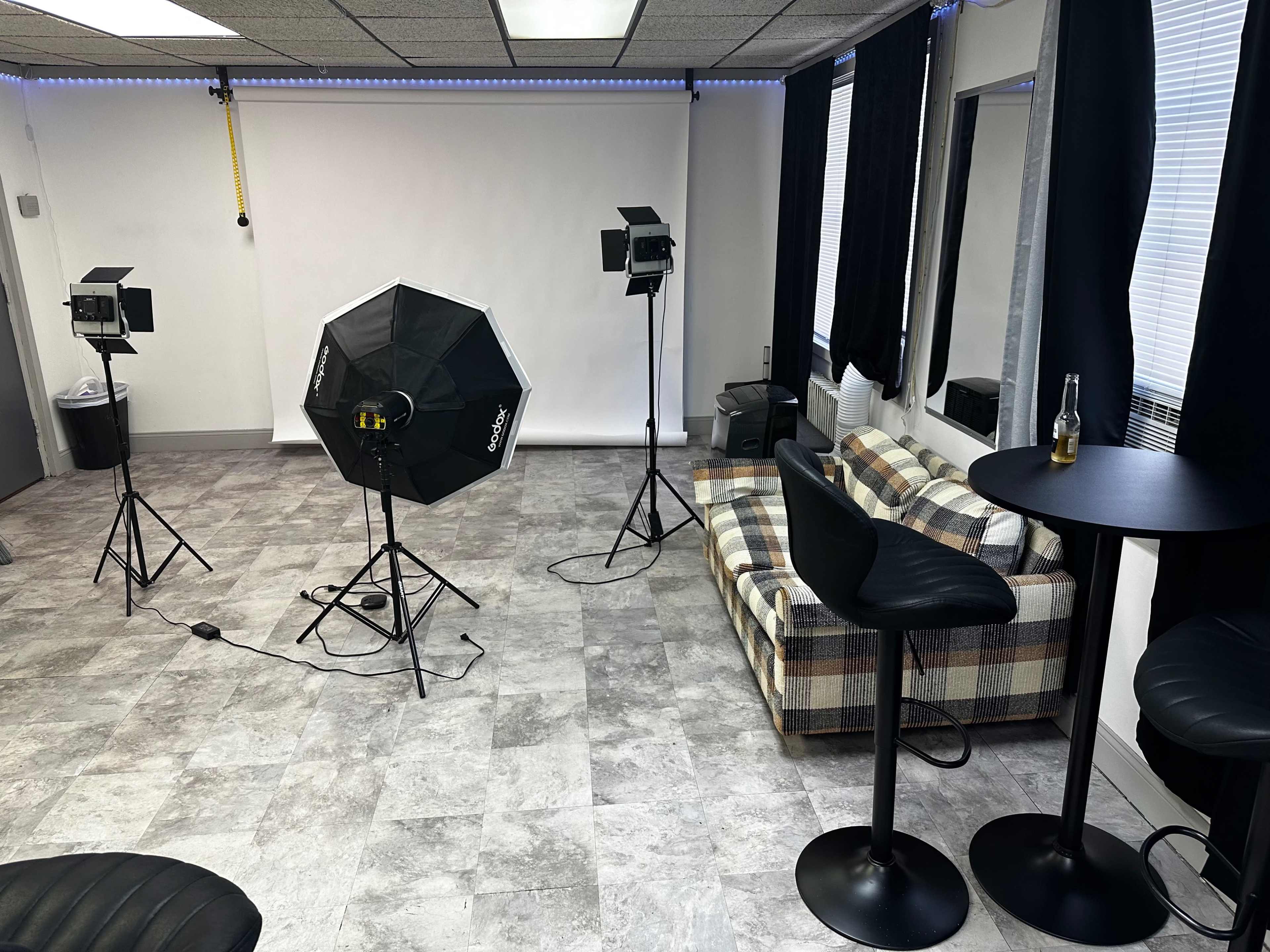 A studio setup with two light stands, an octagonal softbox, a plaid sofa, and a small bar table with stools in a room featuring overhead lighting and a plain backdrop.