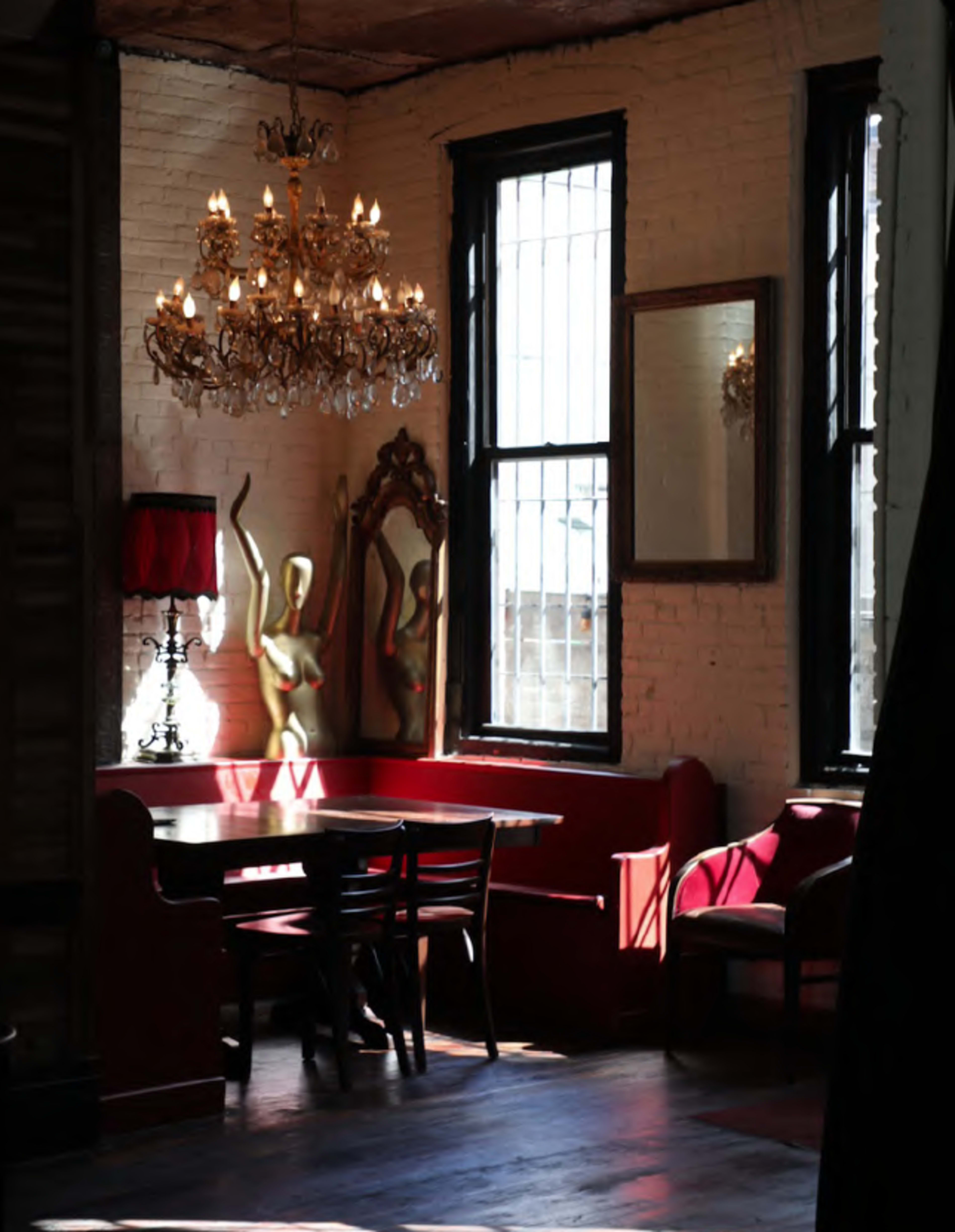 The image shows a well-lit room featuring a chandelier, a red banquette, a wooden table with chairs, and a decorative mirror.