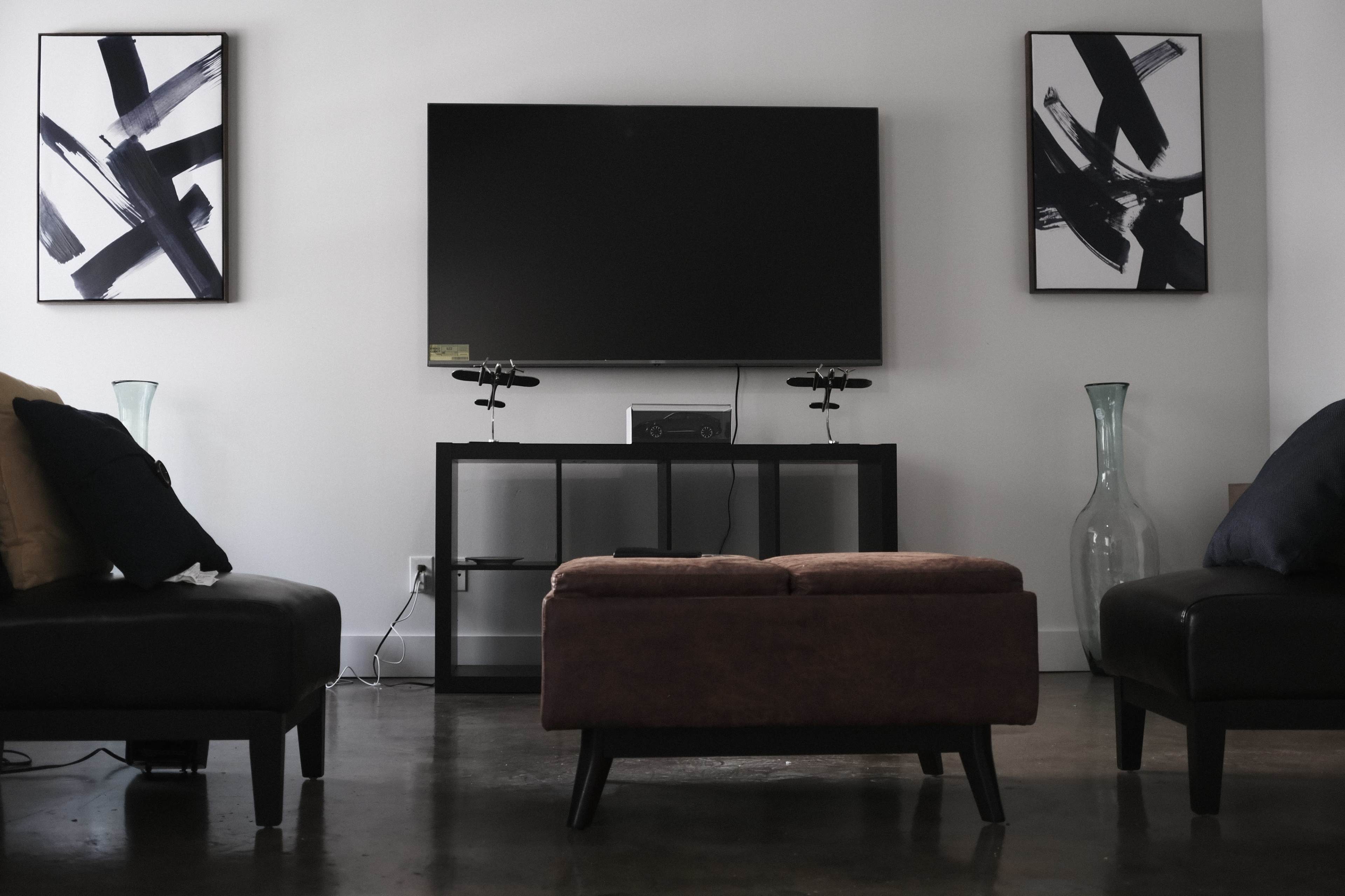 A modern living room features a large flat-screen TV on a dark console table, flanked by abstract black-and-white artwork, with two sofas and an ottoman in front.