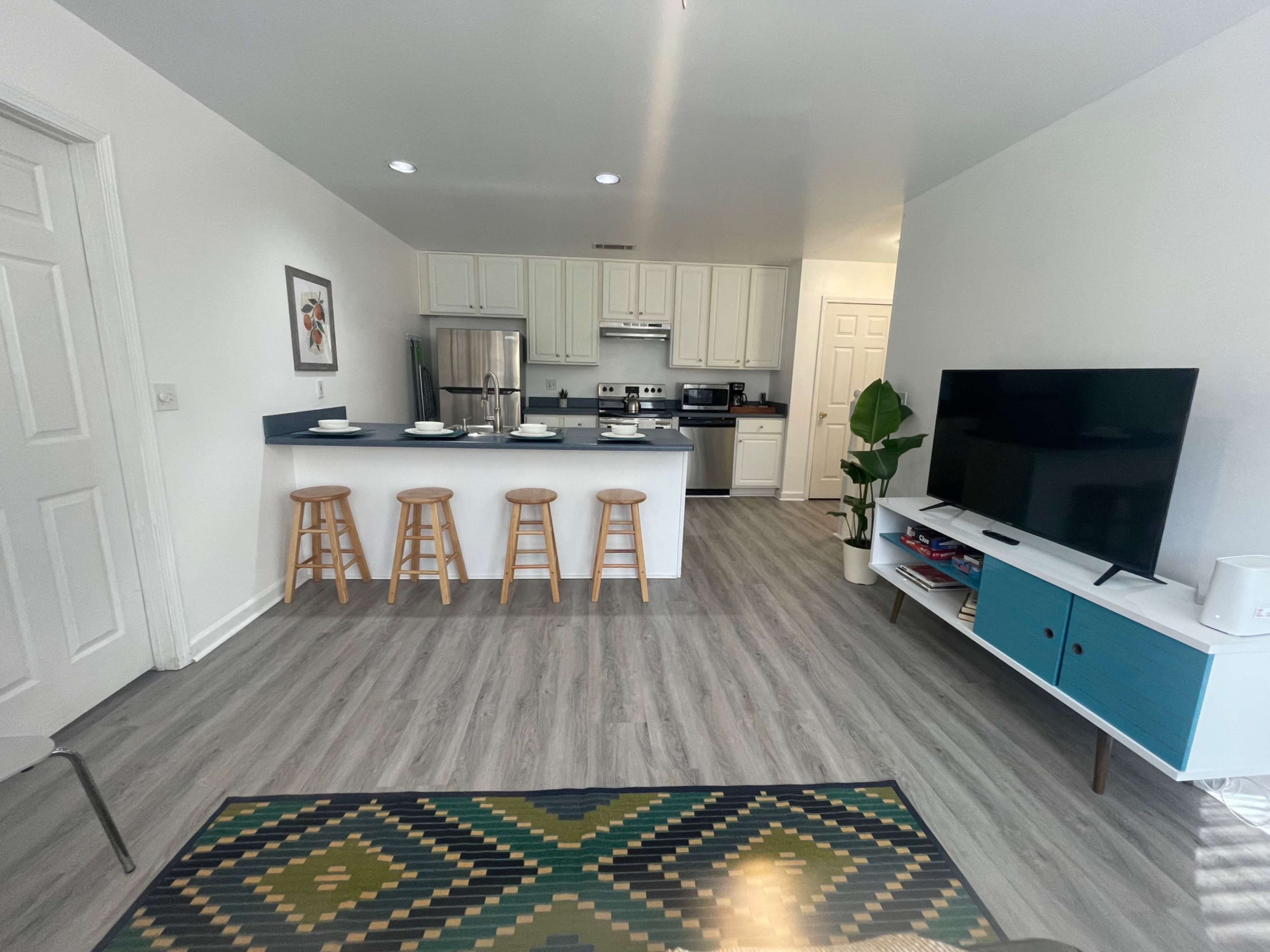 The image shows a modern kitchen and living area with a dining counter, stools, a television stand, and a patterned rug on the floor.