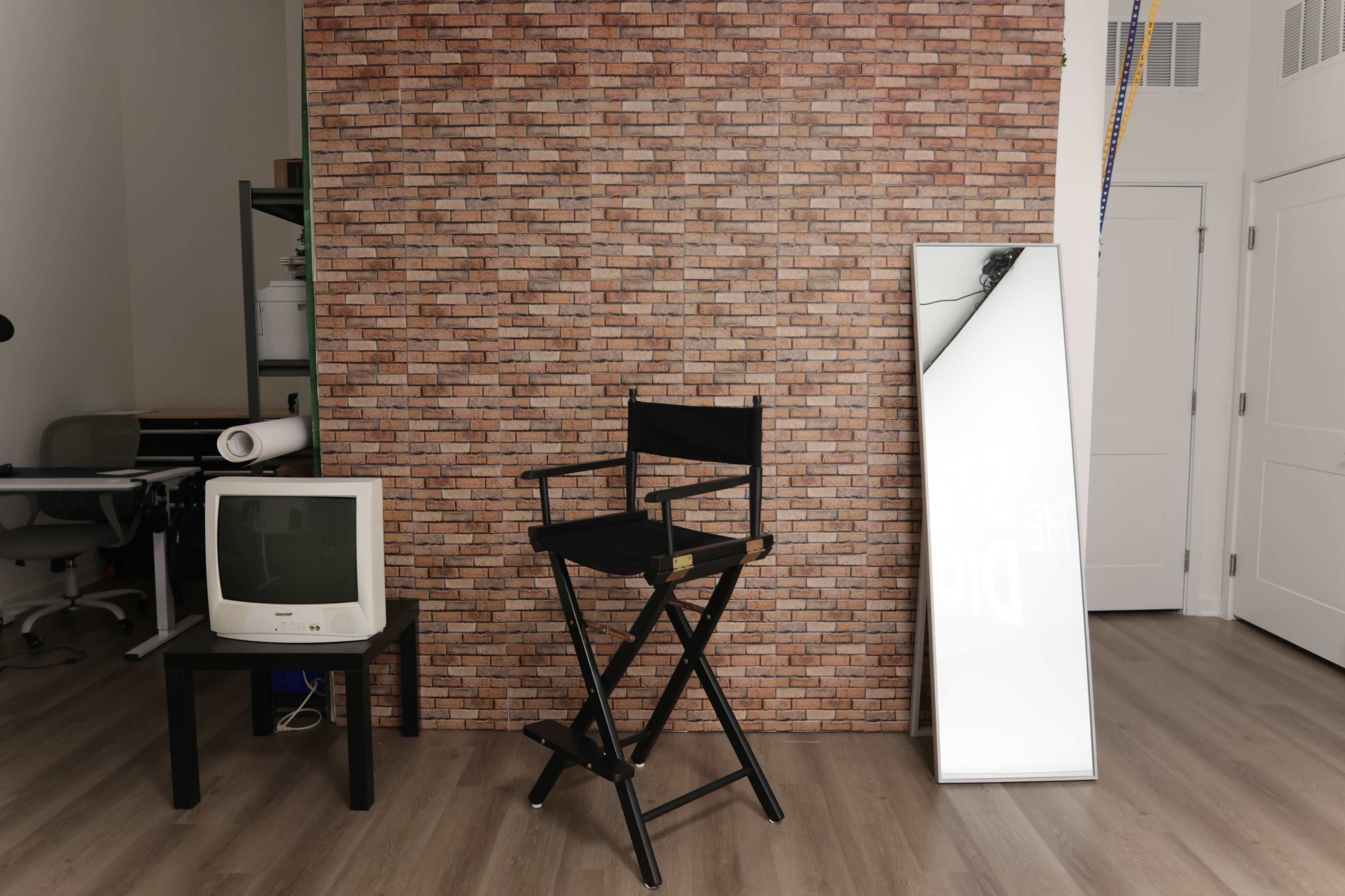 The image shows a black director's chair and an old television set on a small table in front of a brick-patterned wall, with a large mirror leaning against the wall beside them.