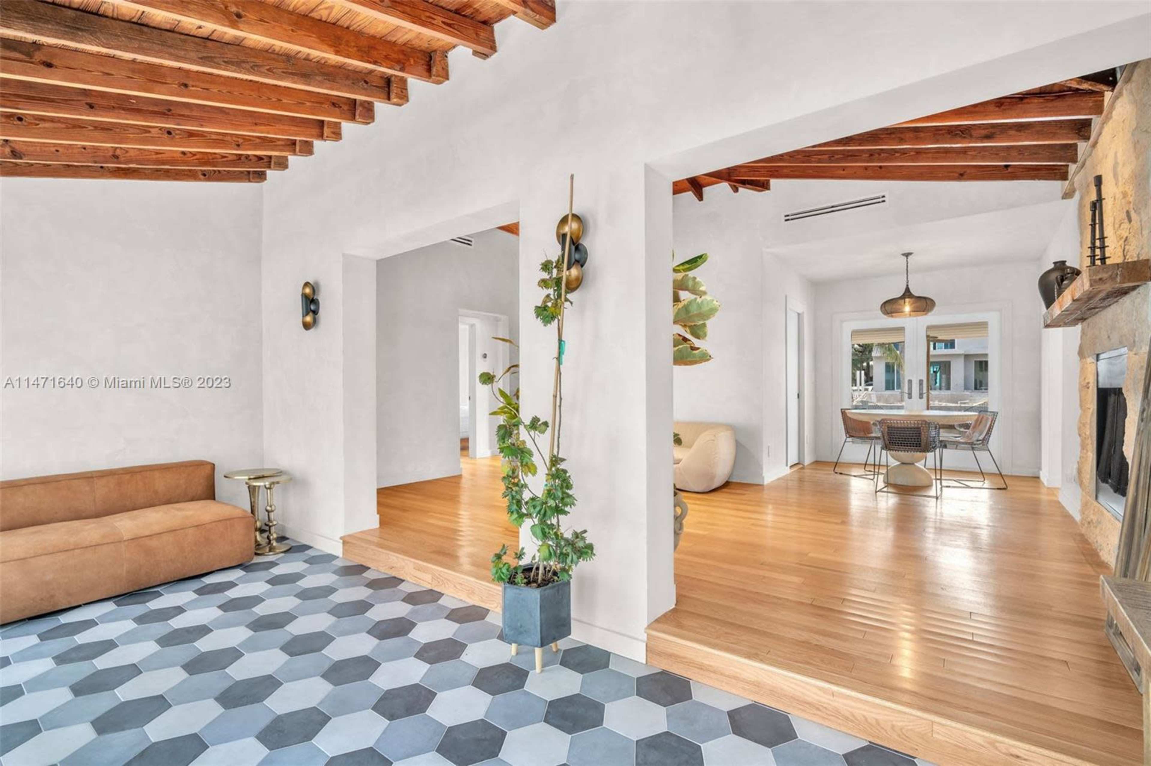 The image shows a modern interior featuring a living area with a sofa, wood beam ceilings, and a two-tone tile floor leading to a dining space.