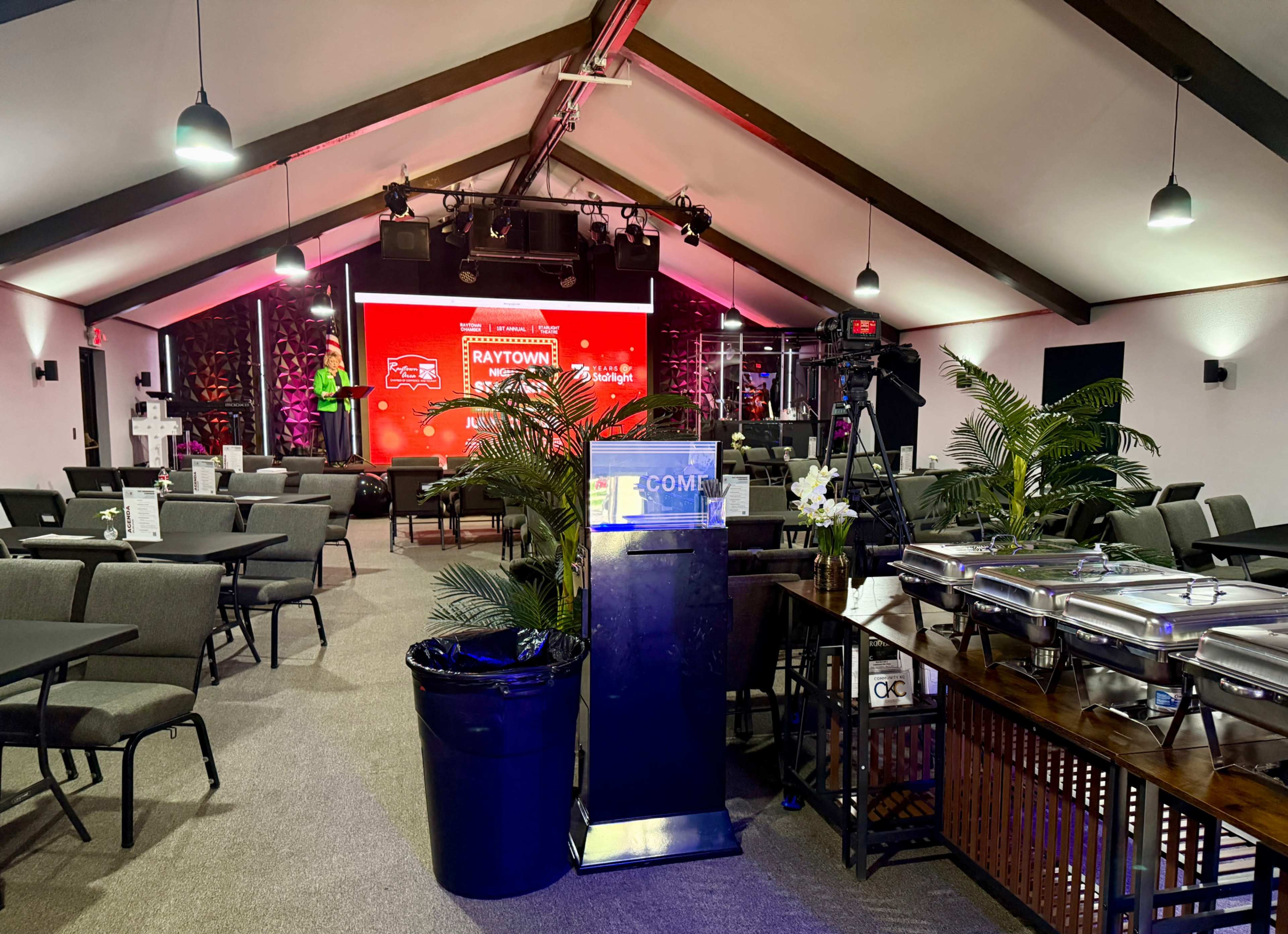 The image shows a spacious indoor venue set up for an event, featuring rows of chairs, a stage with lighting, and food service areas along the side.