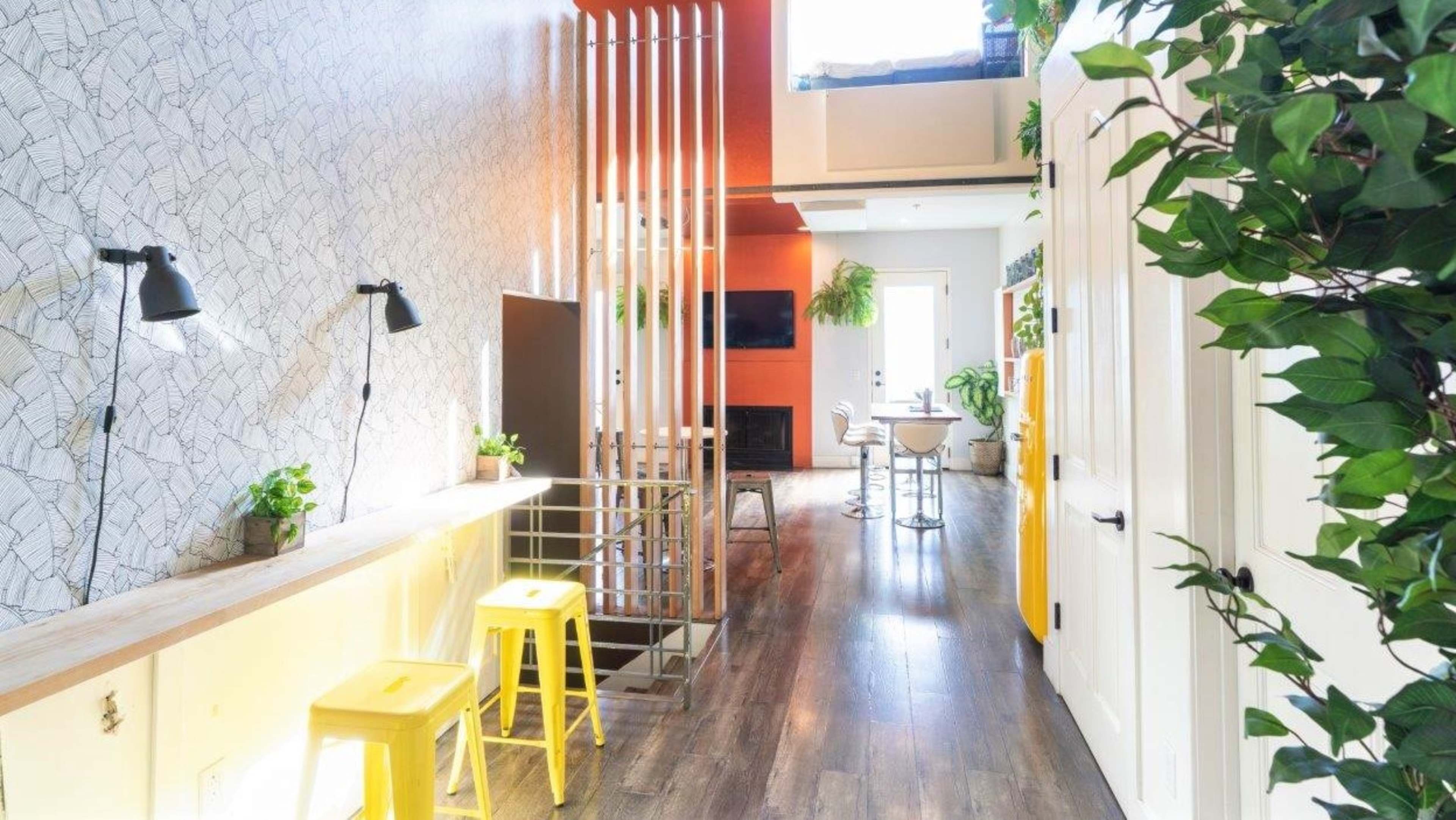 A narrow hallway featuring a combination of wall patterns, a wooden countertop with yellow stools, and greenery along the walls.