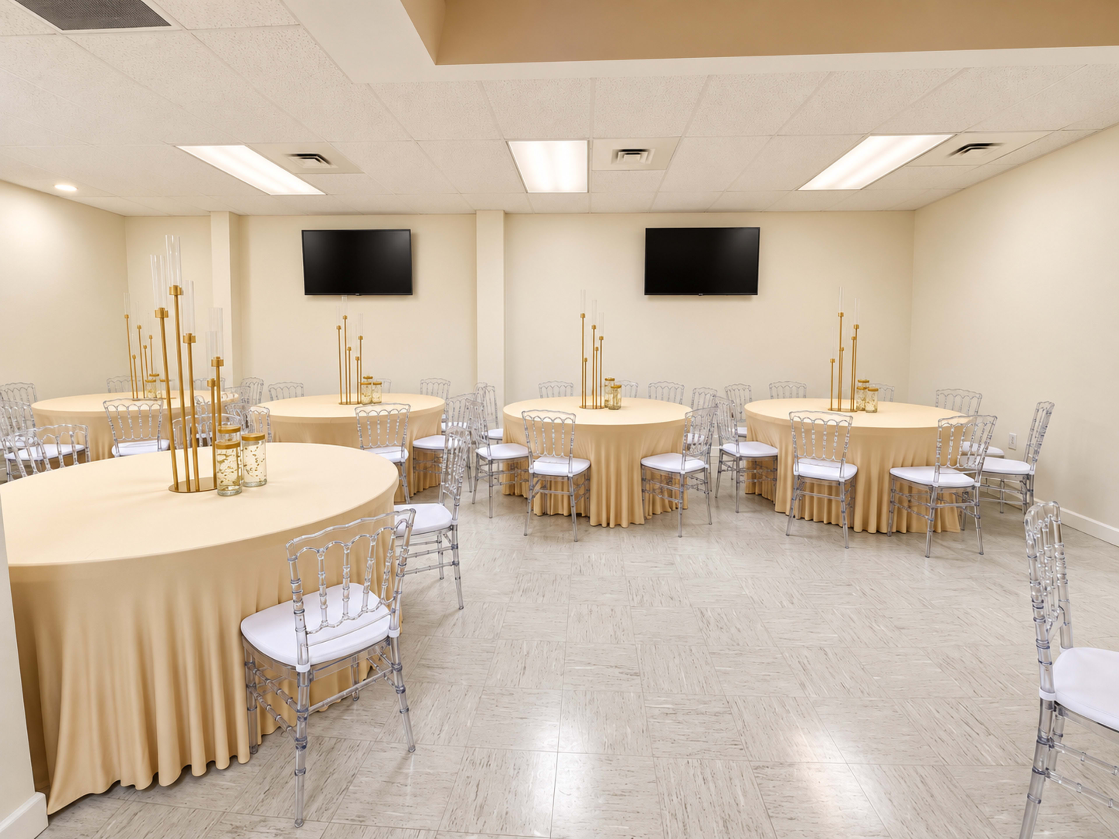 The room features several round tables covered with beige tablecloths, surrounded by clear acrylic chairs, and equipped with two wall-mounted televisions.
