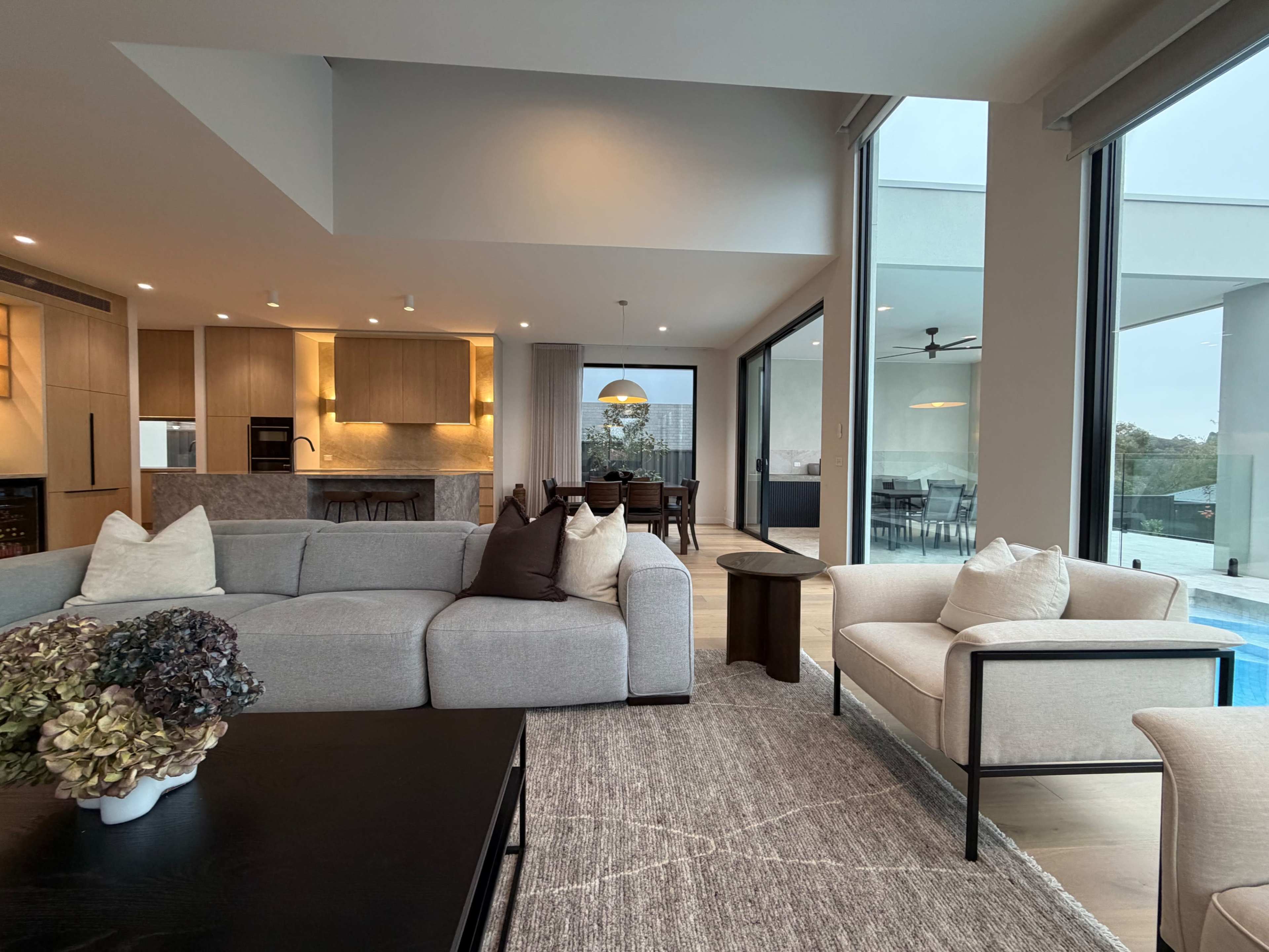 The image shows a modern living area with a light gray sectional sofa, a dark coffee table, and large windows overlooking a pool and outdoor dining space.