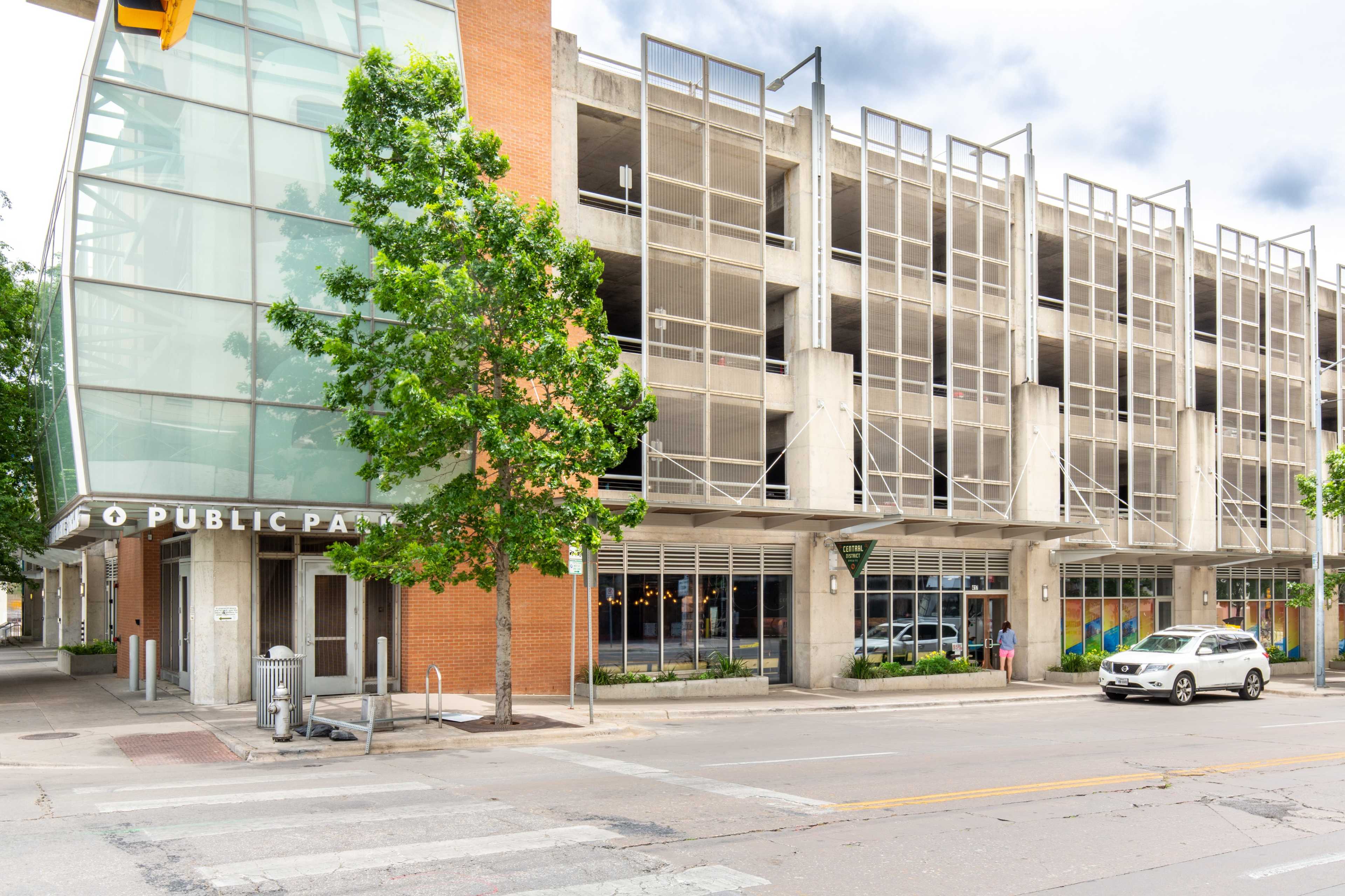 The image shows a modern parking garage building with a glass entrance and colorful murals on the side, situated on a city street.