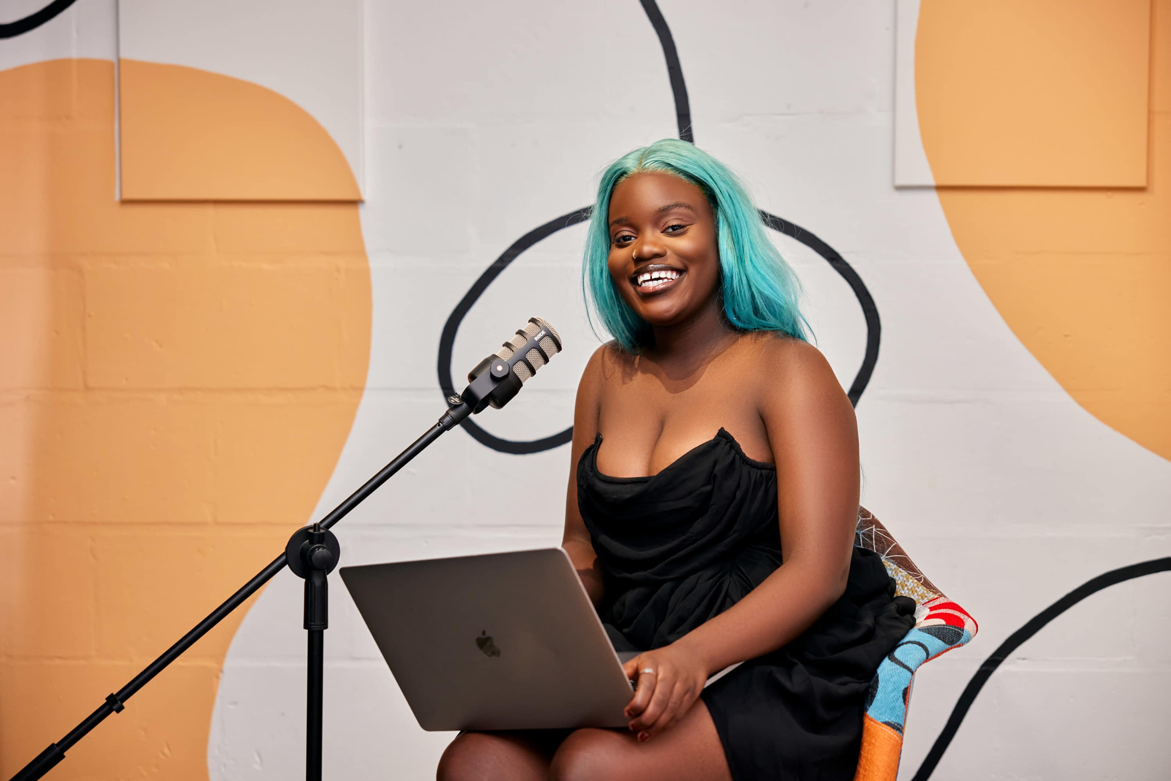 A woman with teal hair sits in front of a microphone and a laptop against a colorful wall.