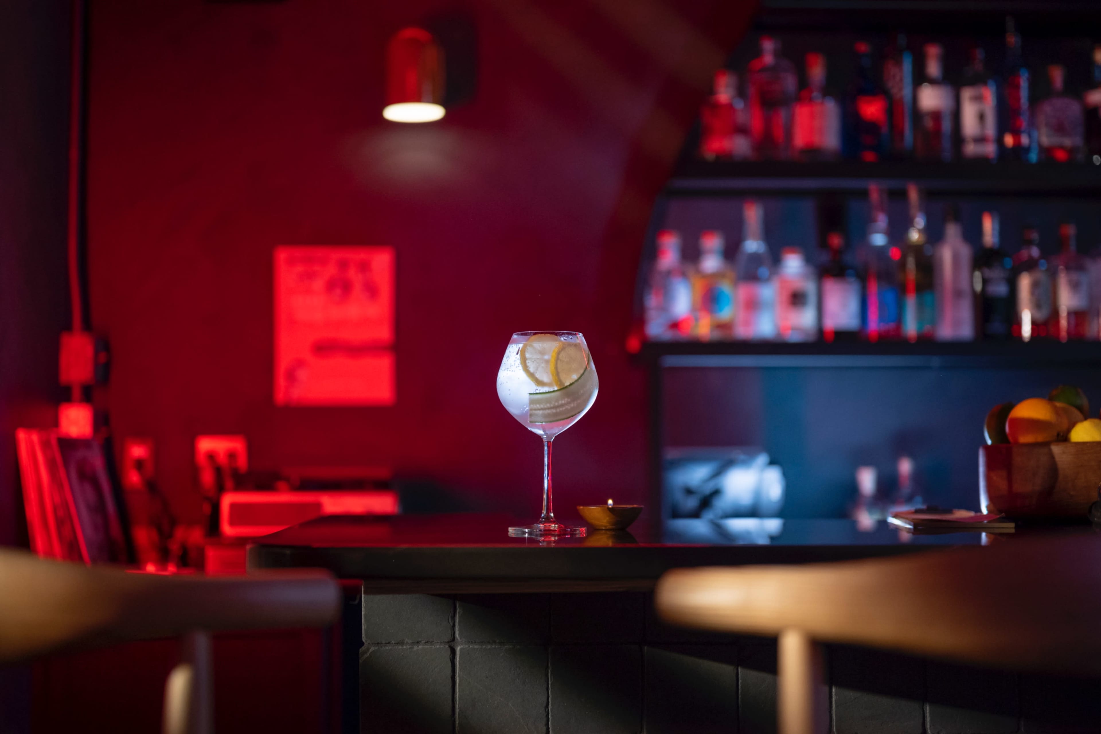 A cocktail glass with a lemon garnish sits on a bar counter, illuminated by red and blue lighting, with bottles visible on a shelf in the background.