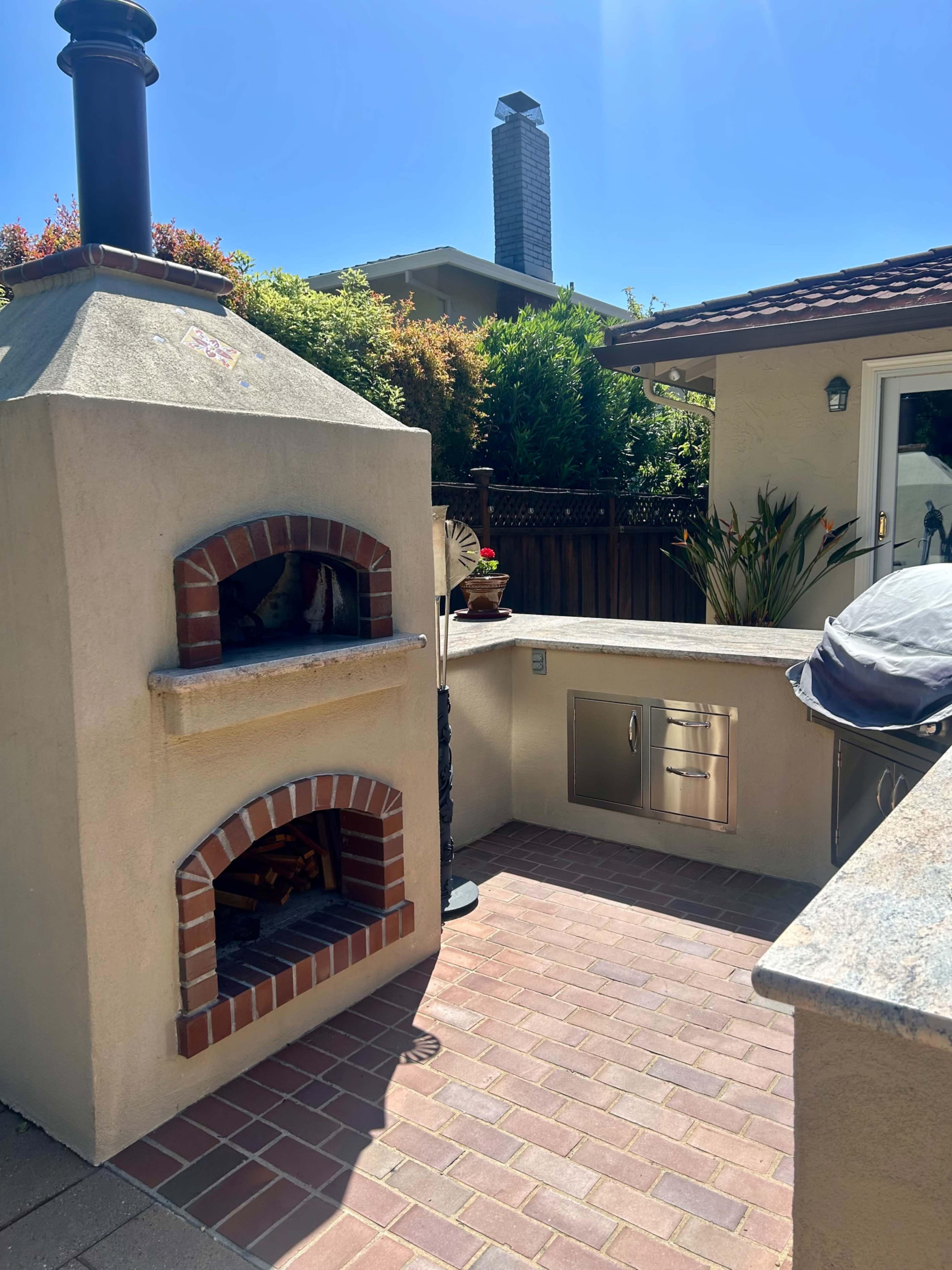 An outdoor space featuring a brick pizza oven next to a built-in grill area, all set on a paved patio.