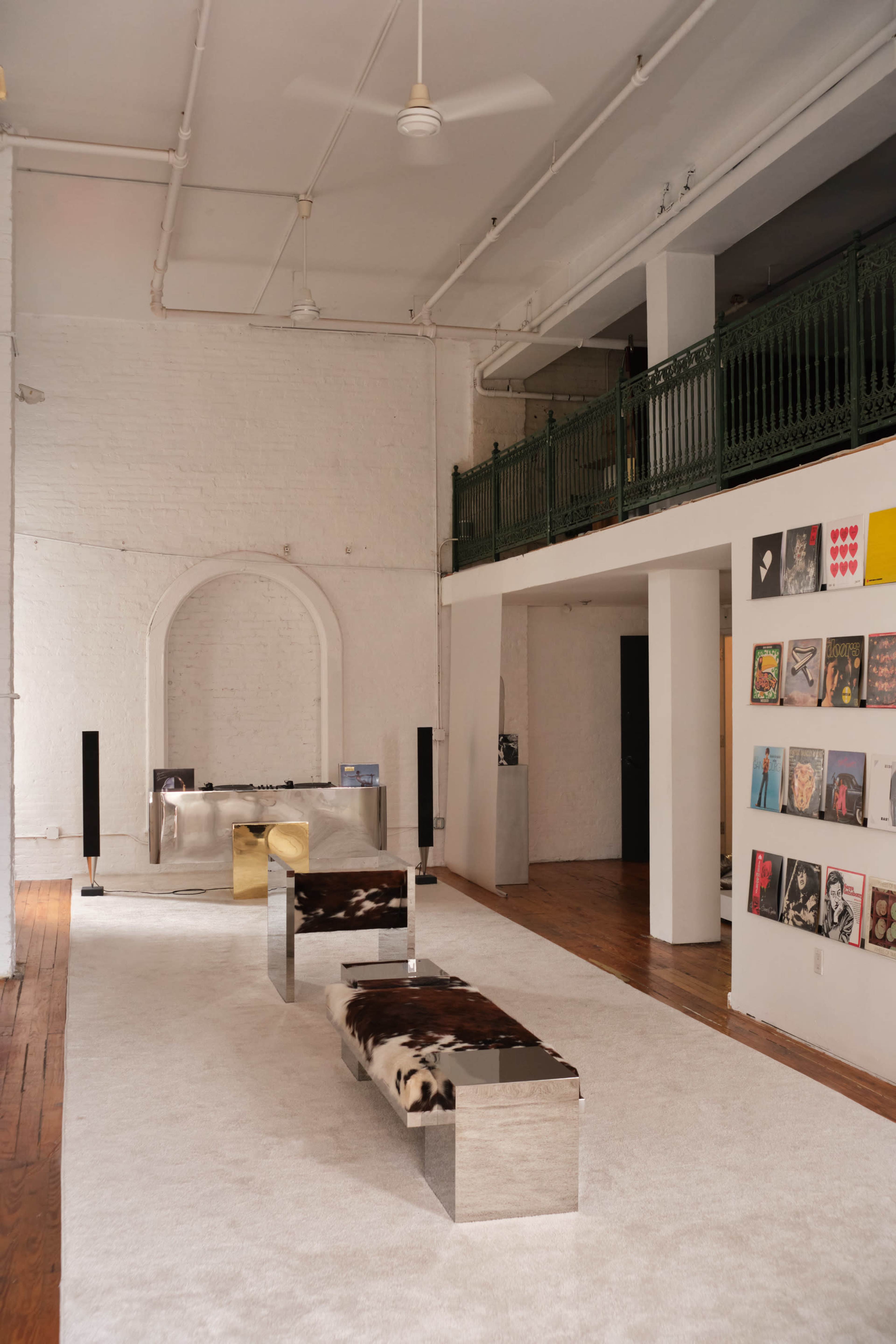 The image depicts a spacious interior featuring a minimalist design with a large area rug, metal benches, a gallery wall of framed artwork, and a second-floor balcony railing.