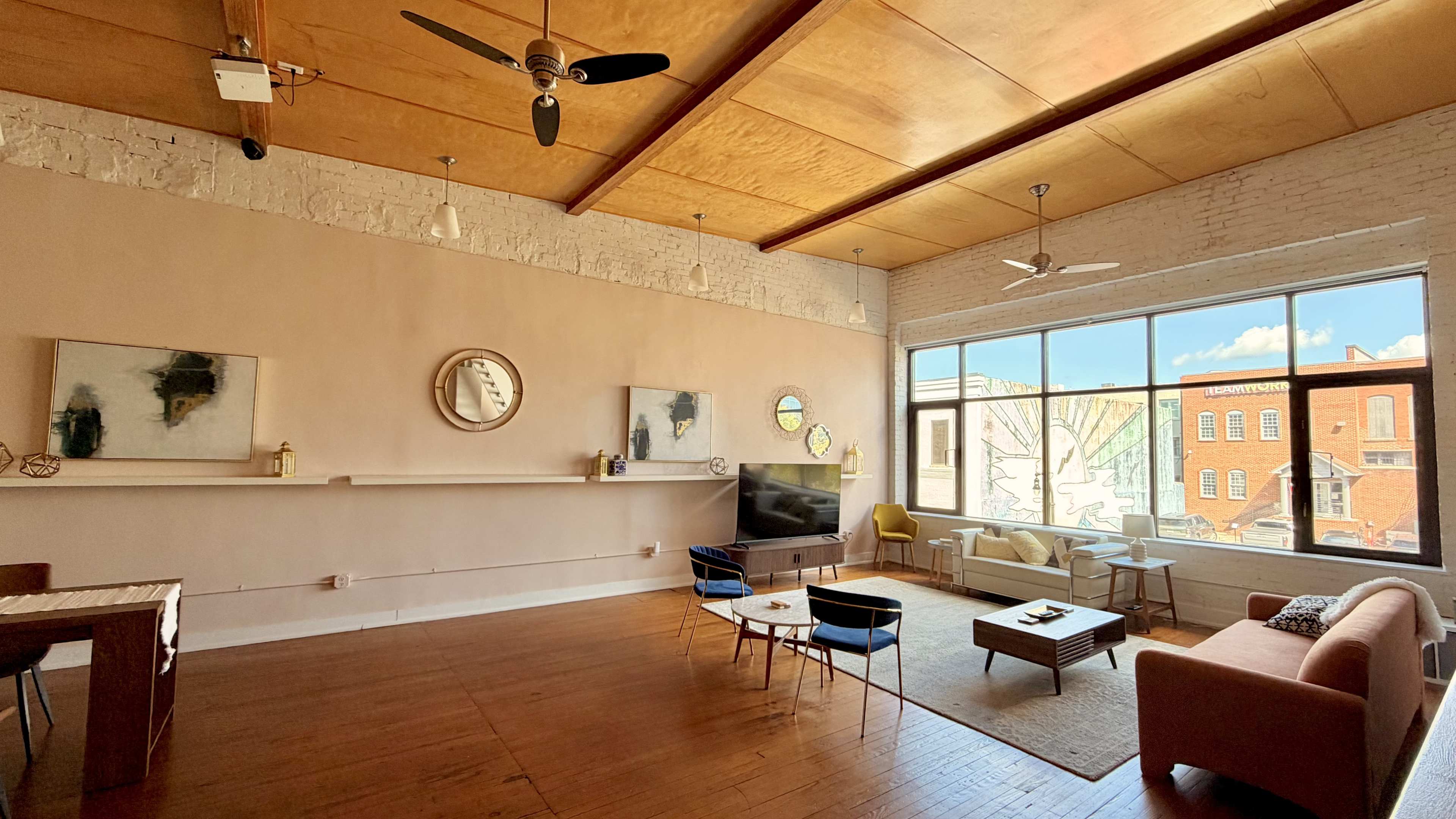 The image depicts a spacious, well-lit living room featuring large windows, wooden ceilings, and a mix of modern furniture.