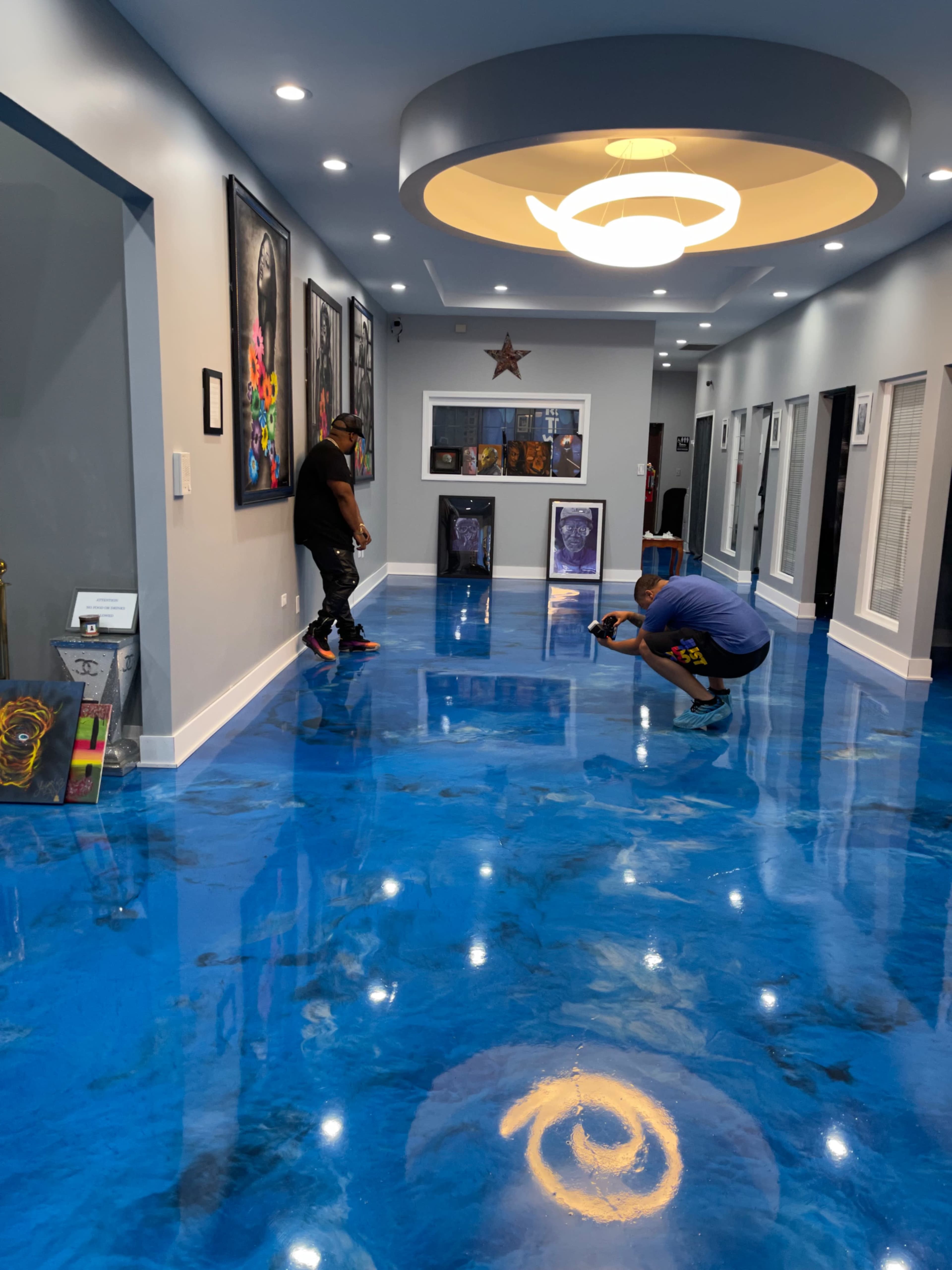Two men are working on a glossy blue floor in a well-lit corridor adorned with artwork and decorative lighting.