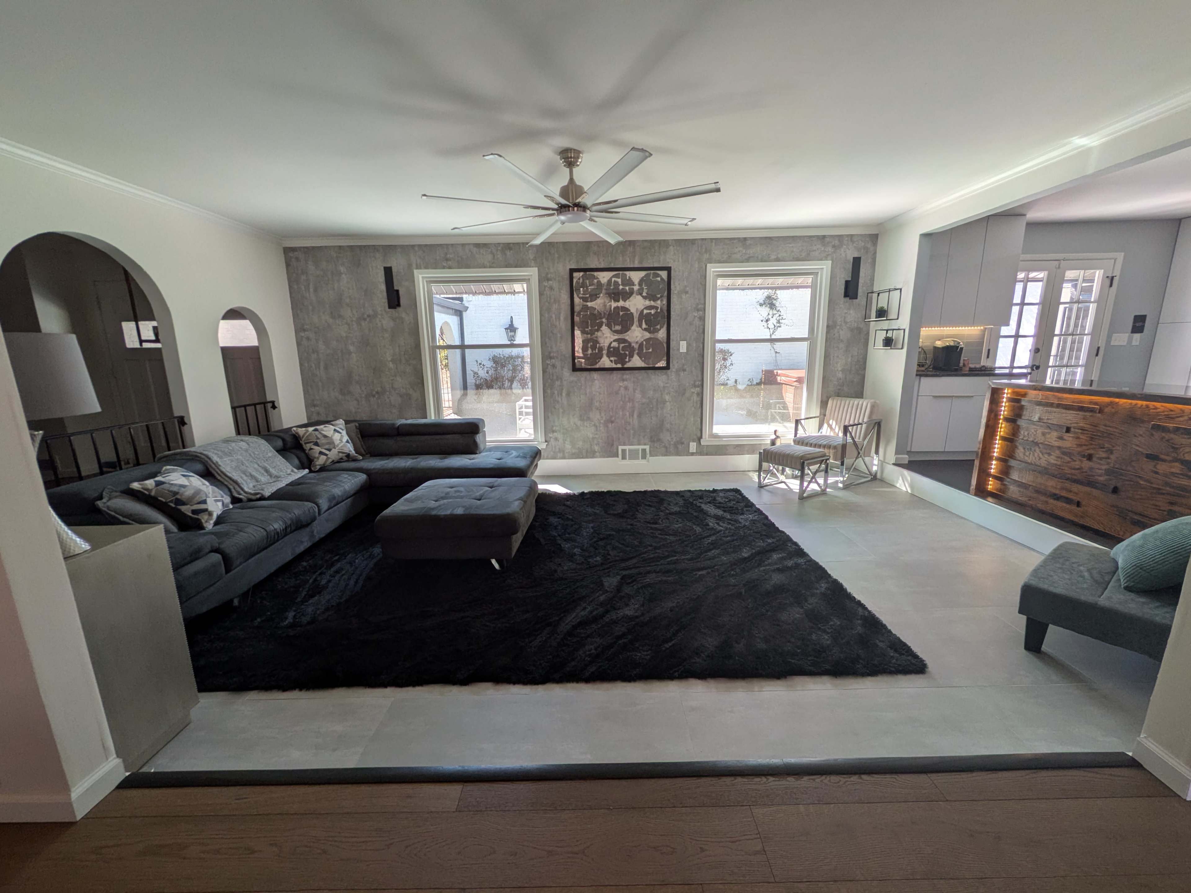The image shows a modern living room with a dark rug, a large sectional sofa, and a kitchen area visible in the background.