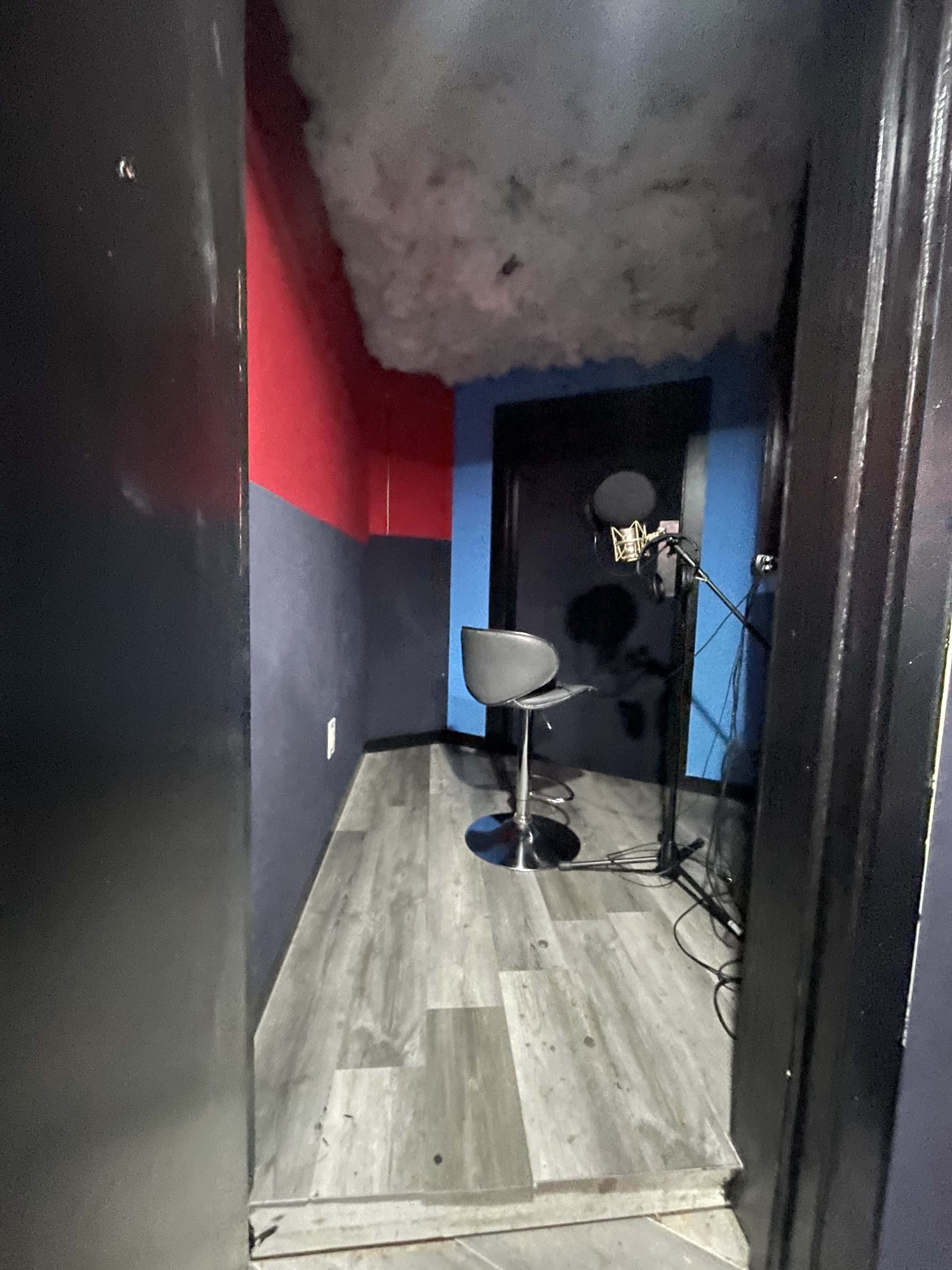 The image shows a small recording booth with a microphone on a stand and a stool, featuring red and blue walls and a textured ceiling.