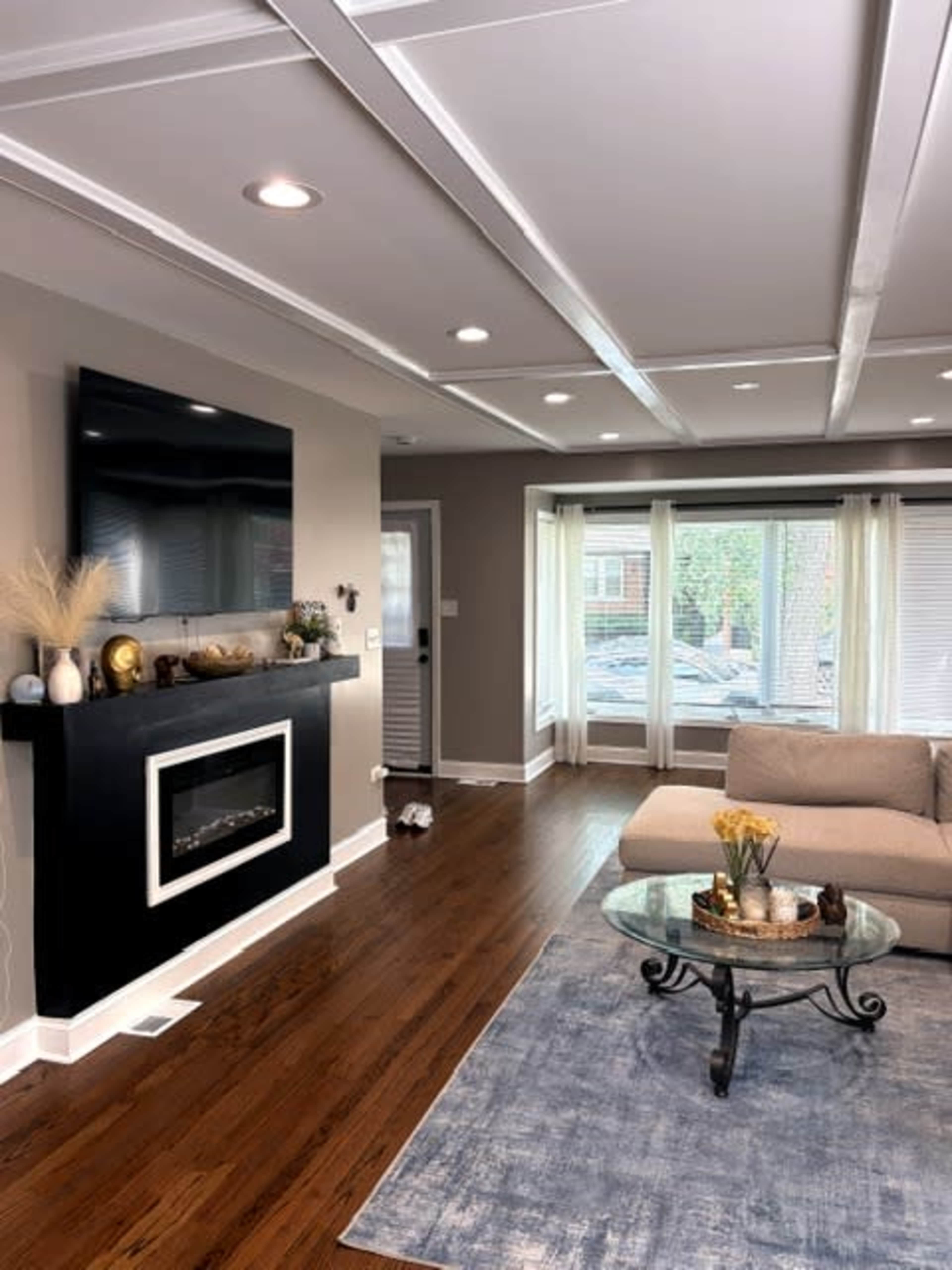 The image shows a contemporary living room with a mounted television, an electric fireplace, a light-colored sofa, and large windows allowing natural light.