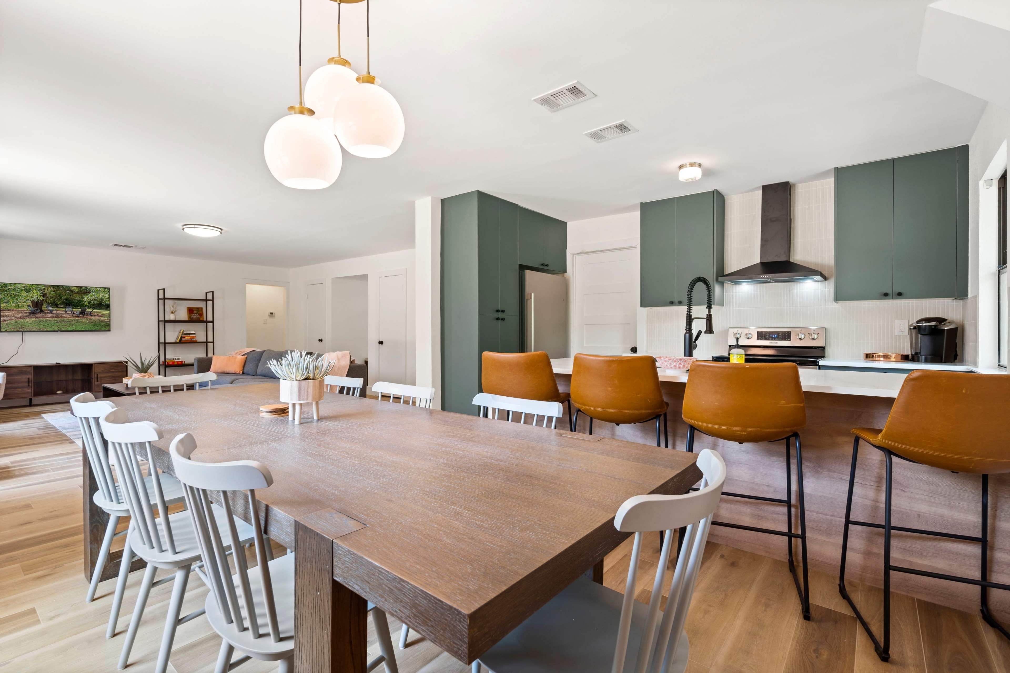 A modern kitchen and dining area featuring a large wooden table with white chairs, a kitchen island with bar stools, and green cabinetry.
