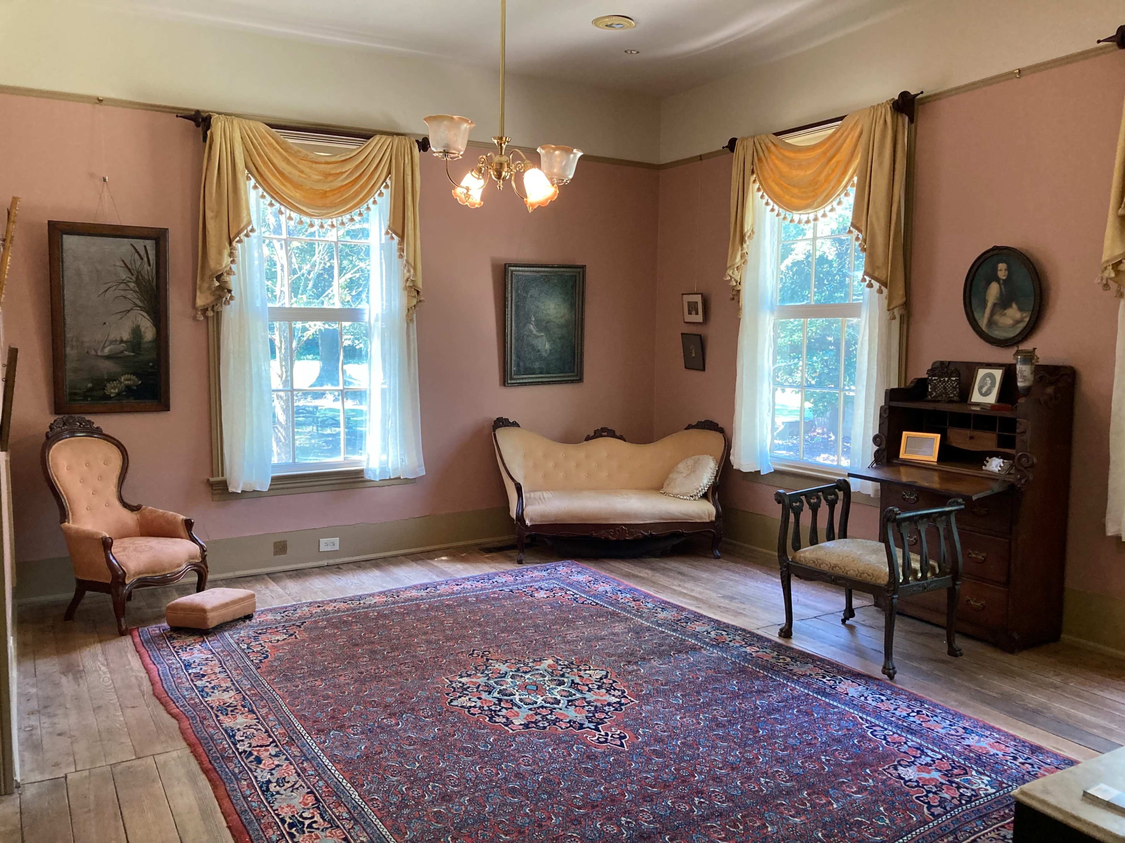 A vintage interior room with a pink wall, two windows dressed in sheer curtains, a patterned rug on the wooden floor, an ornate couch, and a wooden desk and chair.