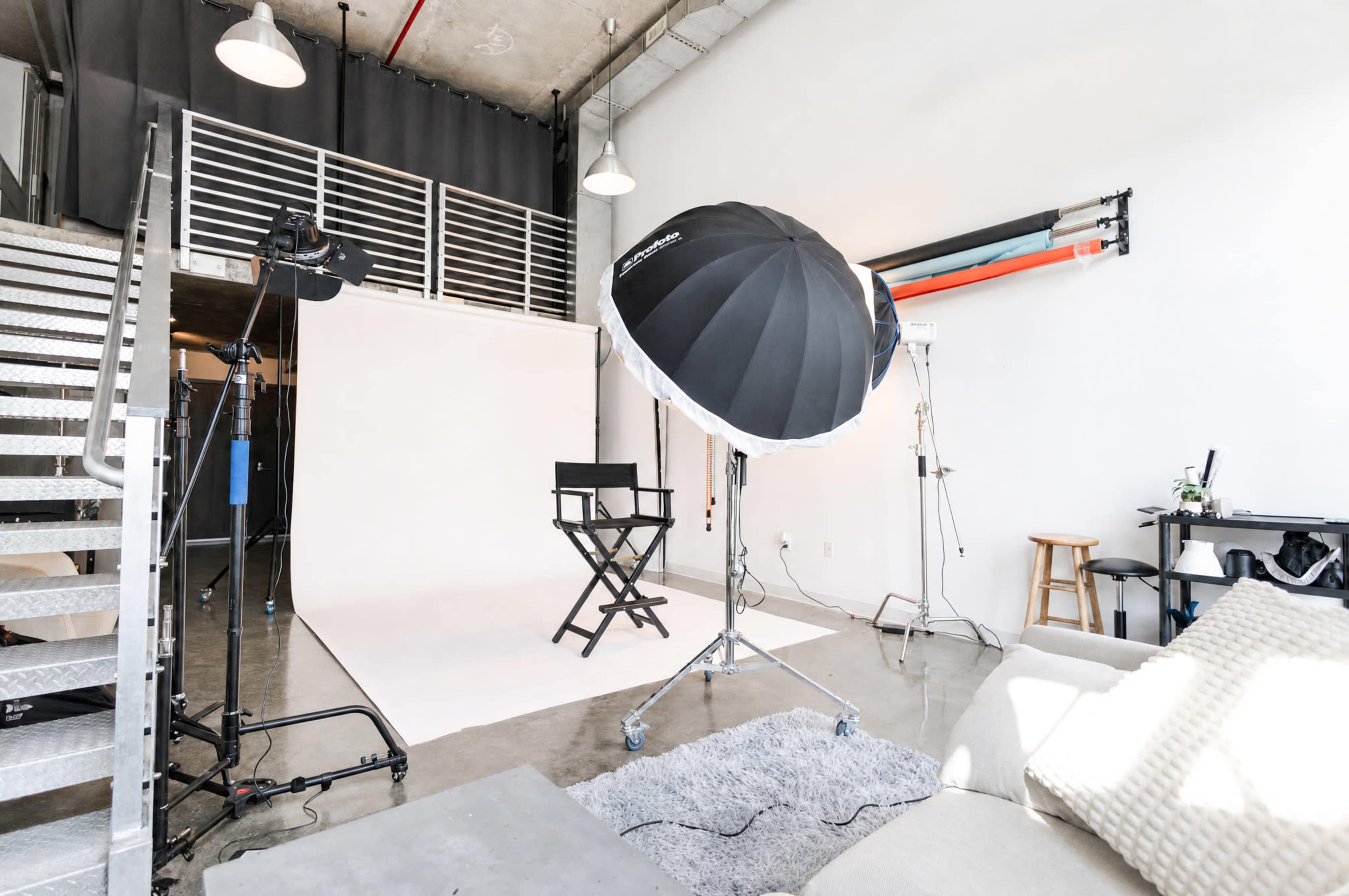 The image shows a photography studio with a large softbox light, a director's chair, a plain backdrop, and various lighting and equipment setups around a modern, spacious interior.