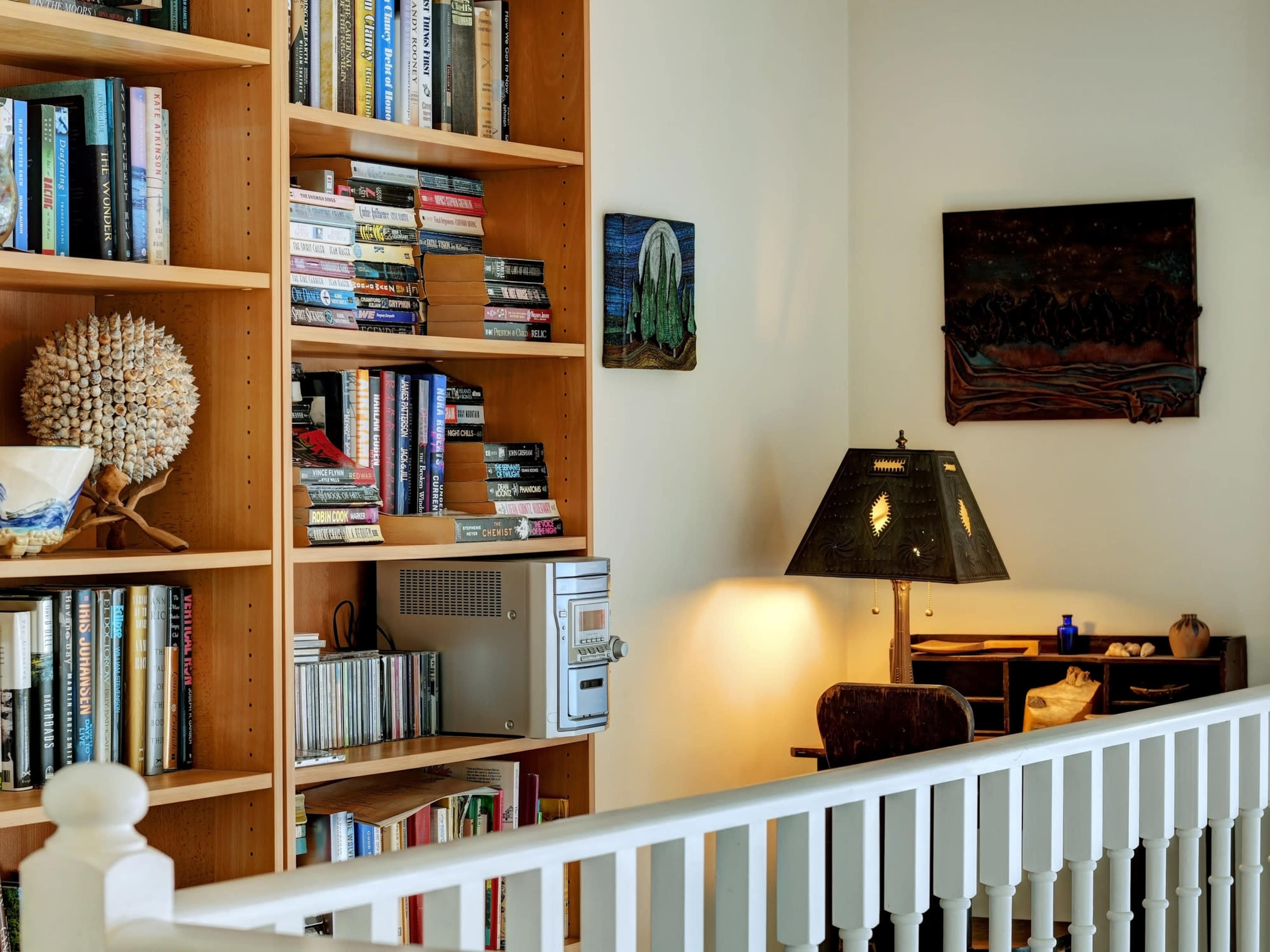 A wooden bookshelf filled with books and DVDs stands next to a lamp and a small desk in a room with light-colored walls.