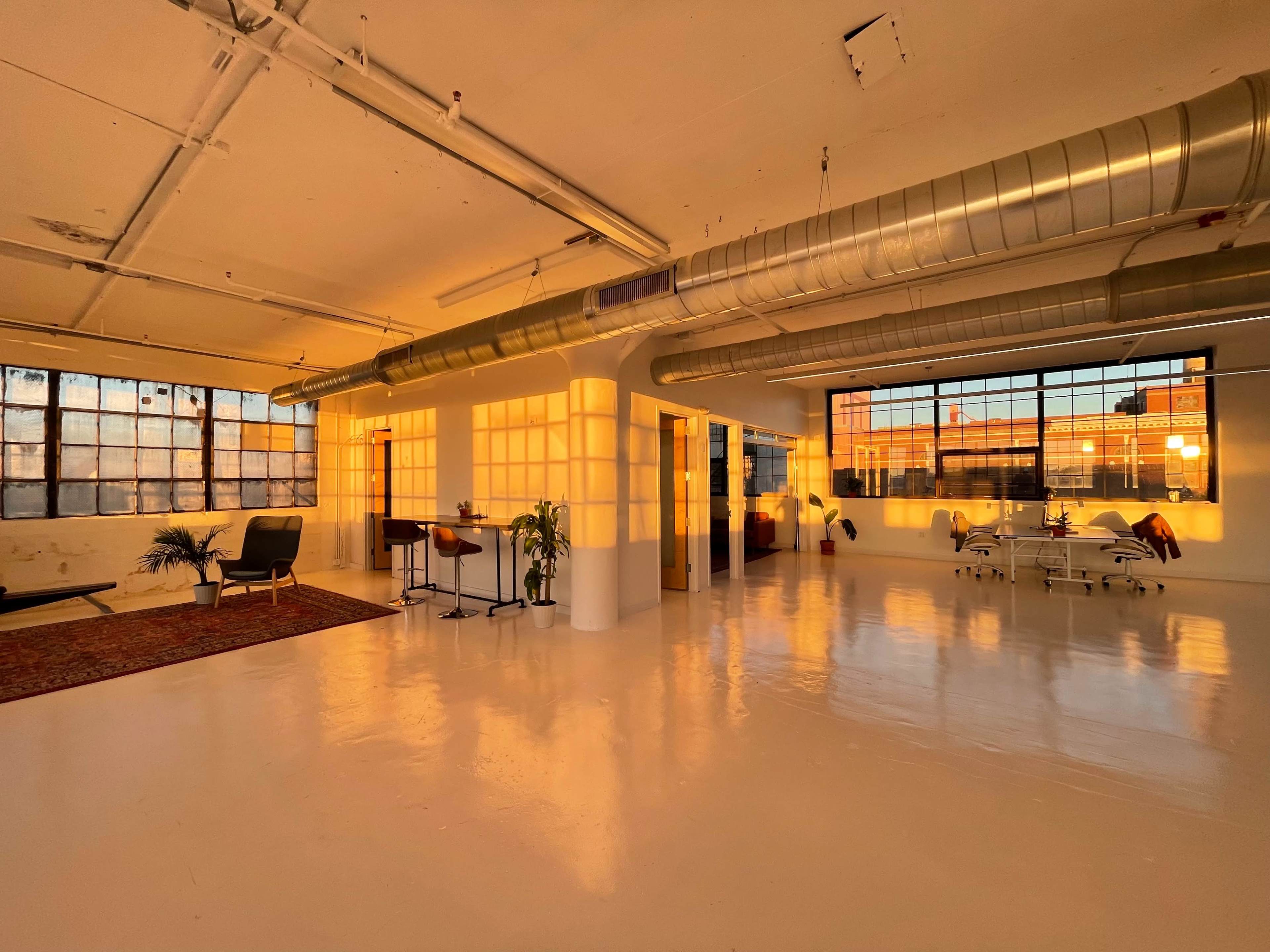 A spacious, well-lit industrial studio features large windows, polished white floors, and minimalist furnishings under soft sunset lighting.