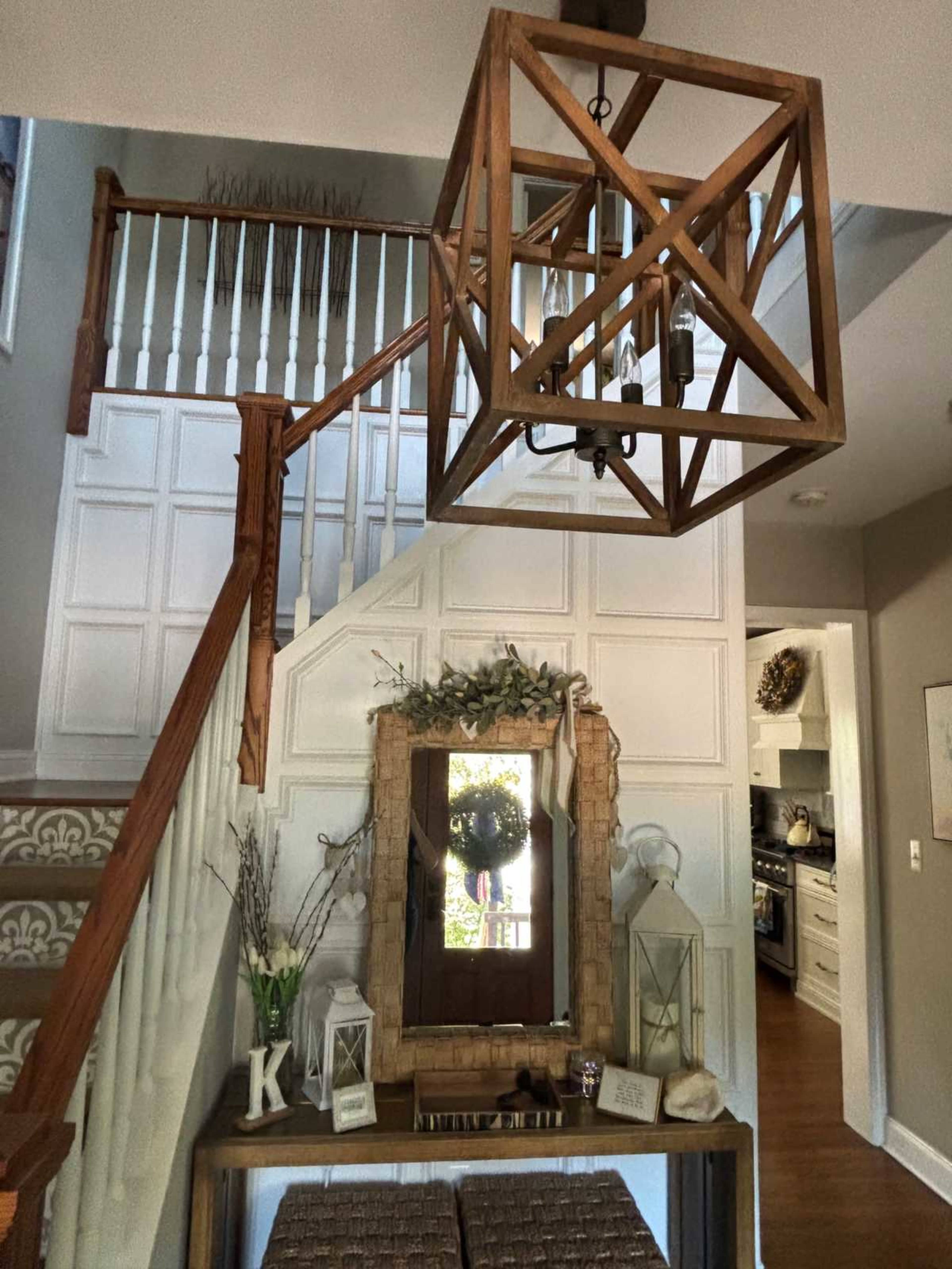 The image shows a well-decorated entryway with a wooden chandelier, staircase, and a table featuring a mirror and decorative items.