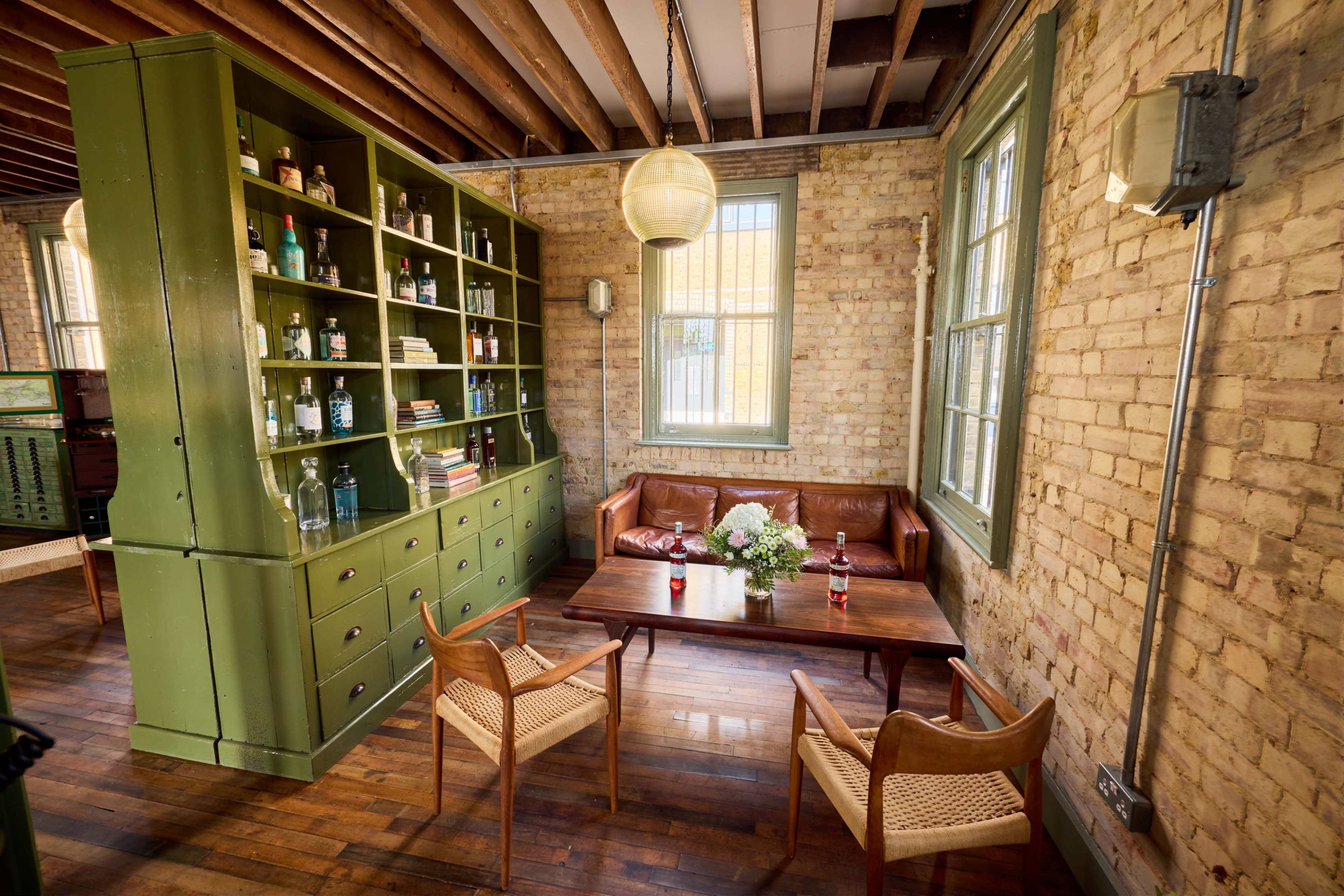 The image shows a cozy living space featuring a green bookshelf, a brown leather couch, a wooden table, and two chairs, all set against a backdrop of exposed brick walls and wooden beams.