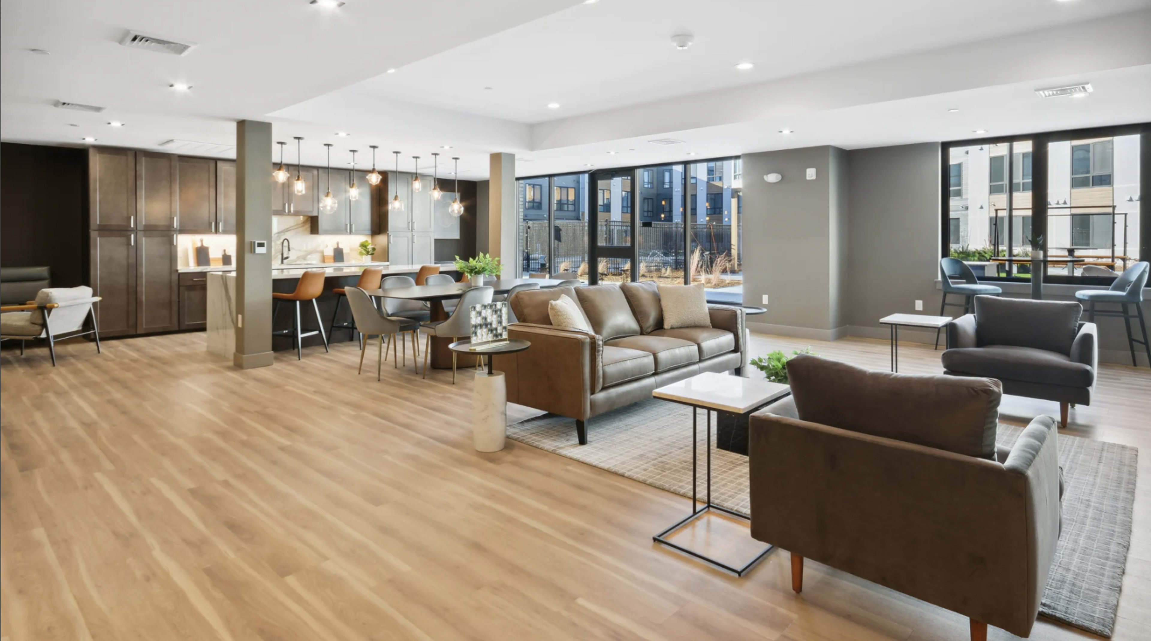 The image shows a modern communal living space featuring a kitchen, dining area, and seating arrangements with large windows allowing natural light.