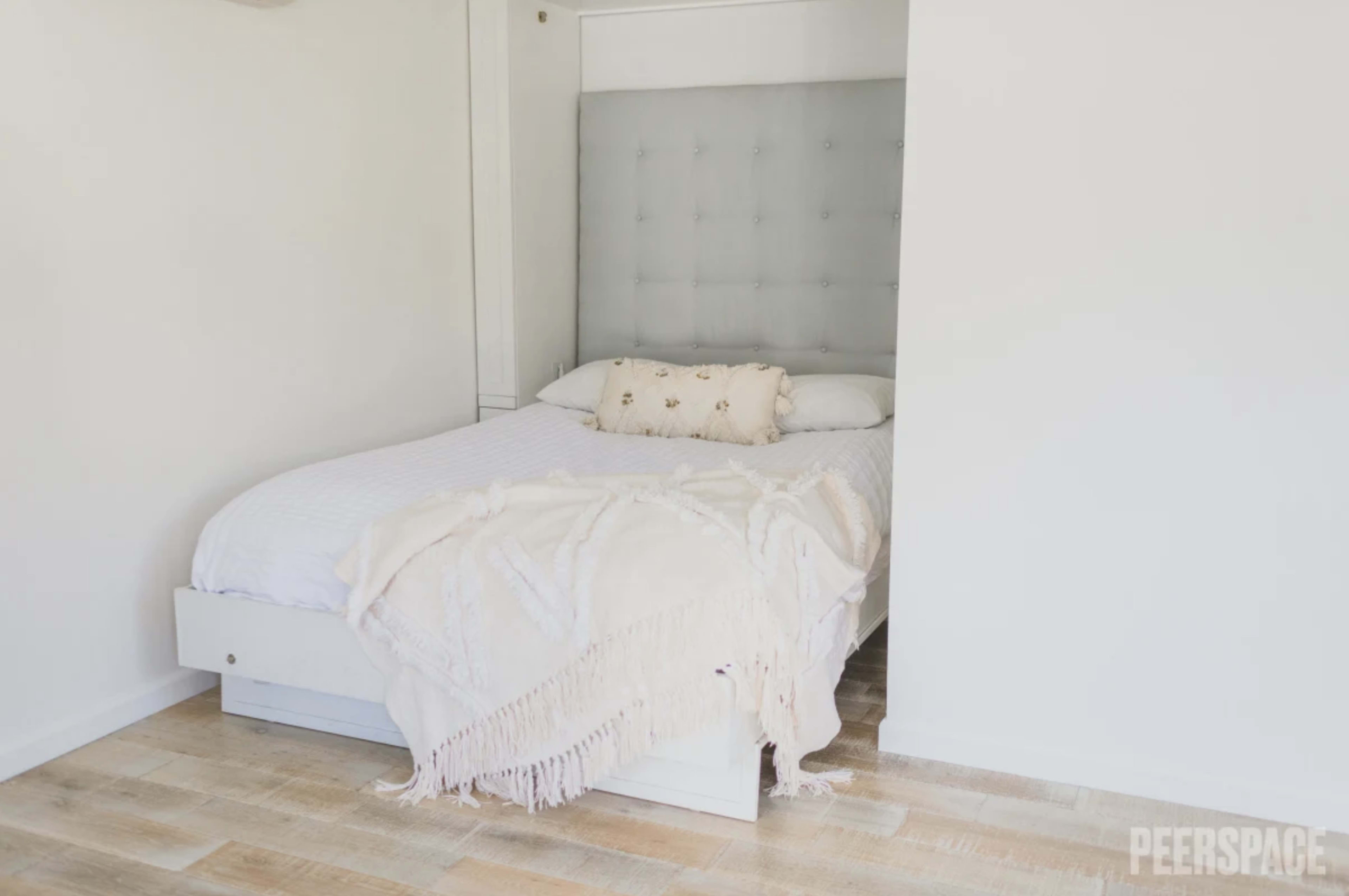A neatly made bed with a decorative pillow and a fringed blanket is positioned against a light-colored wall in a bedroom.