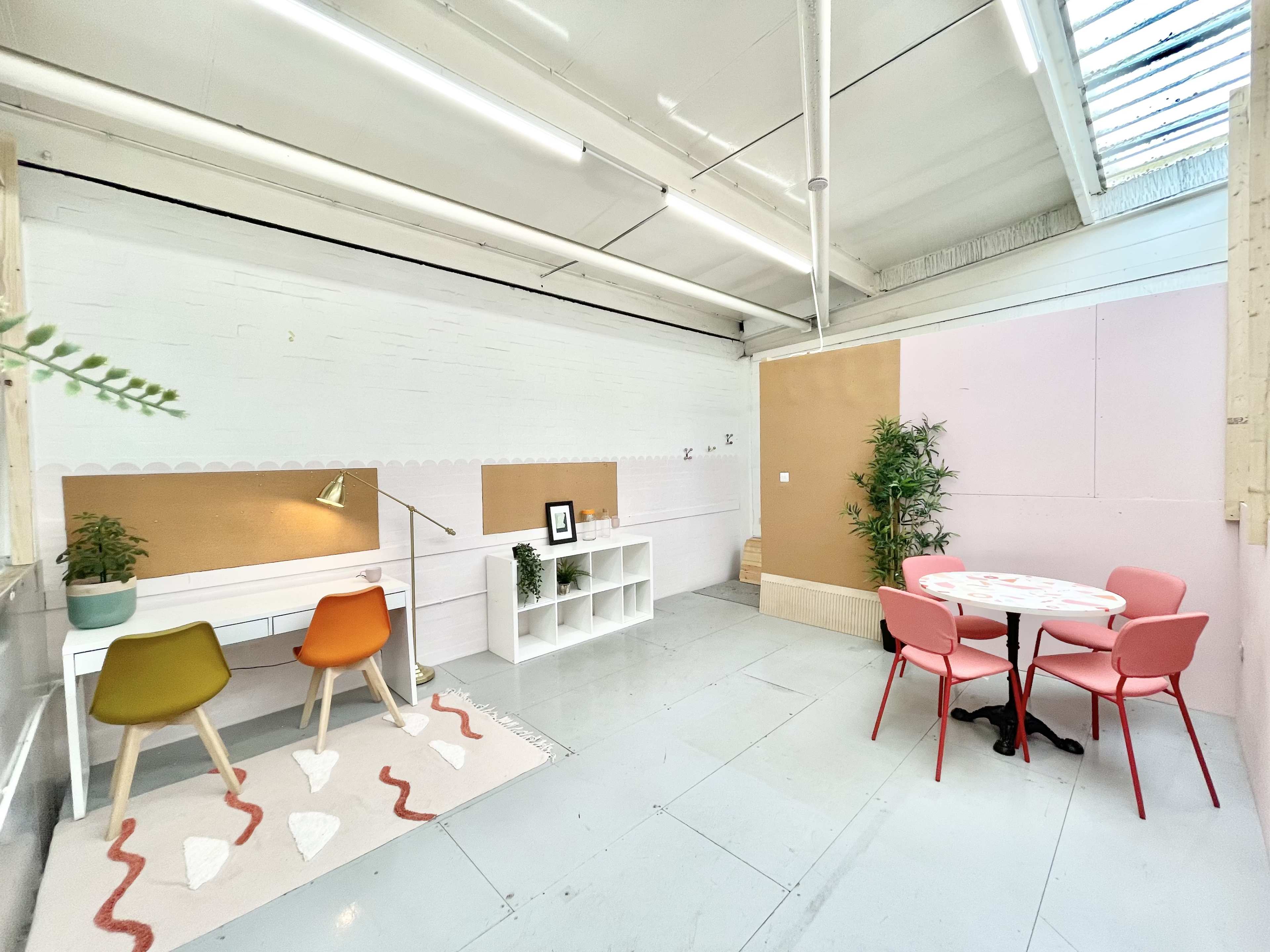 A bright, minimalist workspace featuring a desk, colorful chairs, a small table, and decorative plants in a light-colored room.