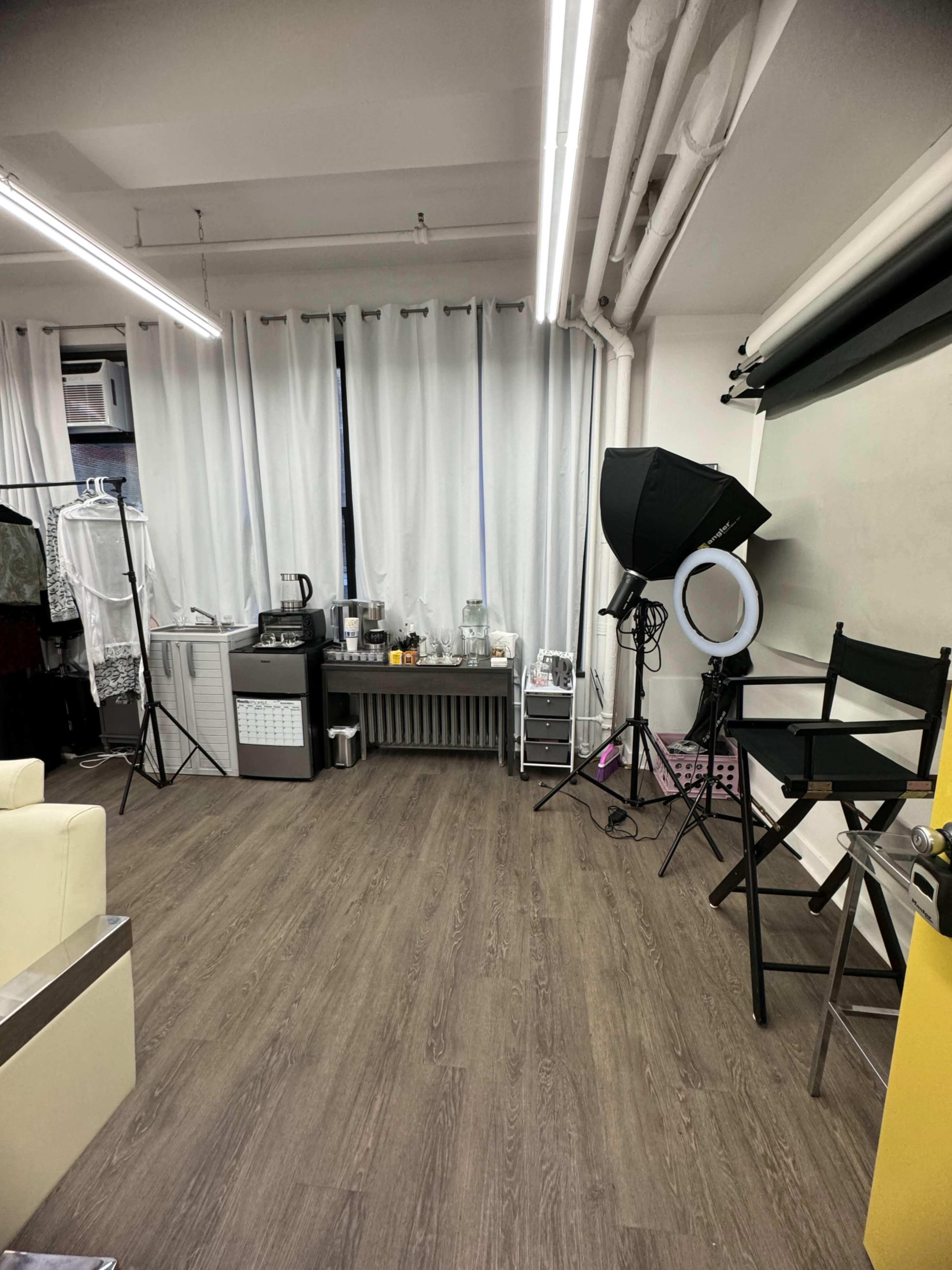 A spacious room with a wooden floor, featuring white curtains, a kitchenette area, a lighting setup, and a black director's chair.