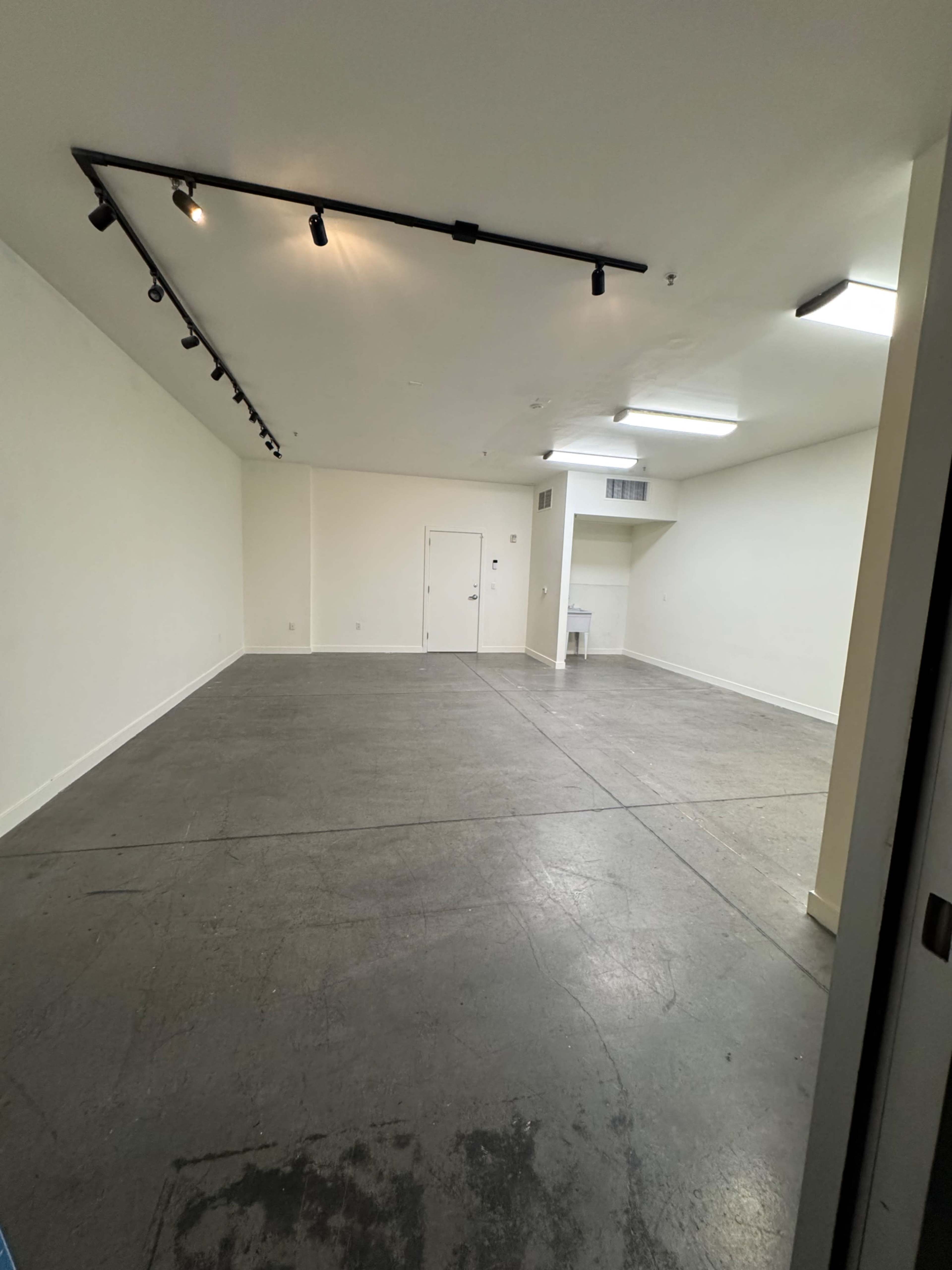 The image shows an empty room with concrete flooring and white walls, featuring track lighting along the ceiling.