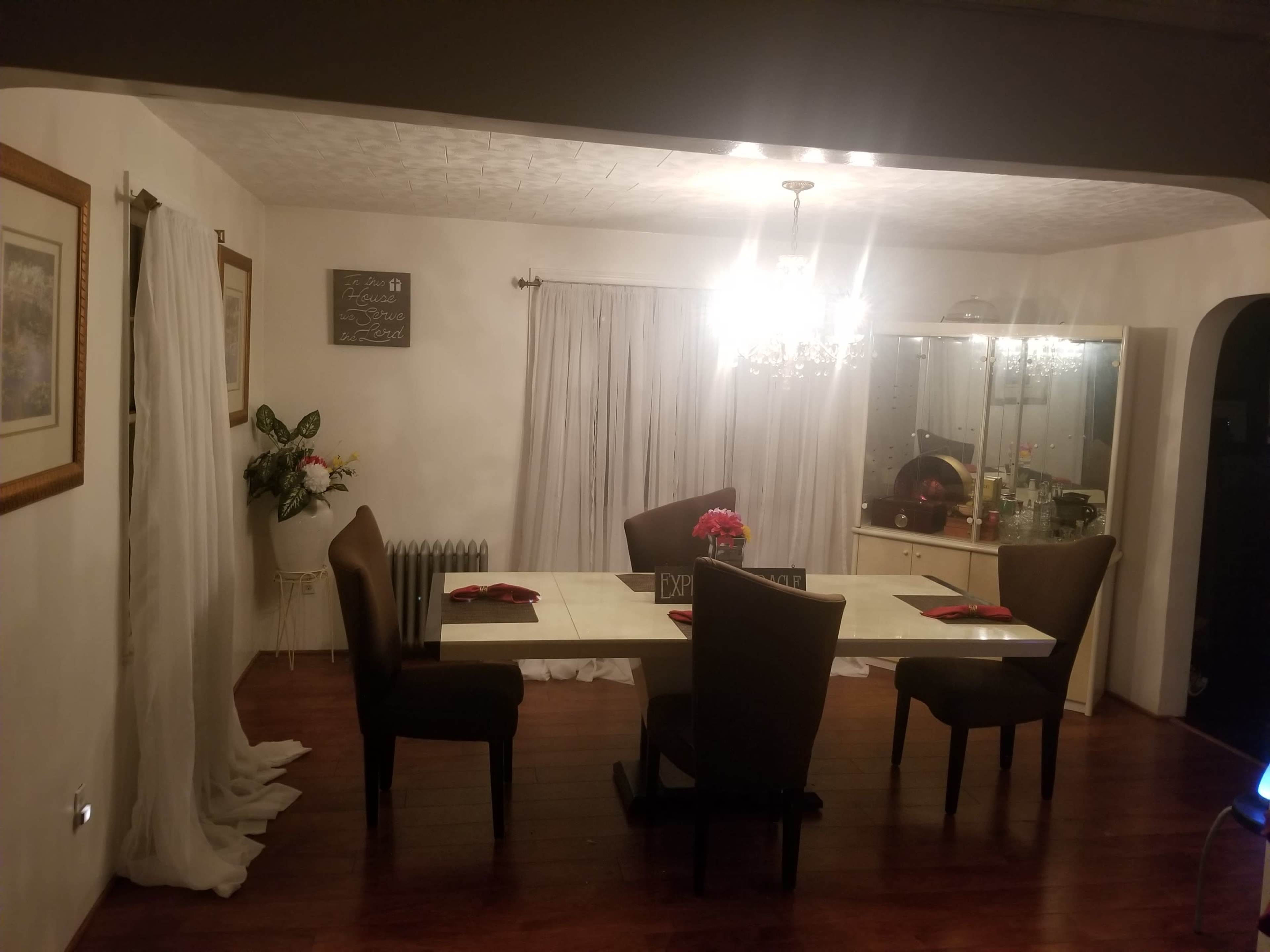 A dining room features a white table surrounded by four dark chairs, with a chandelier above and a cabinet in the background.