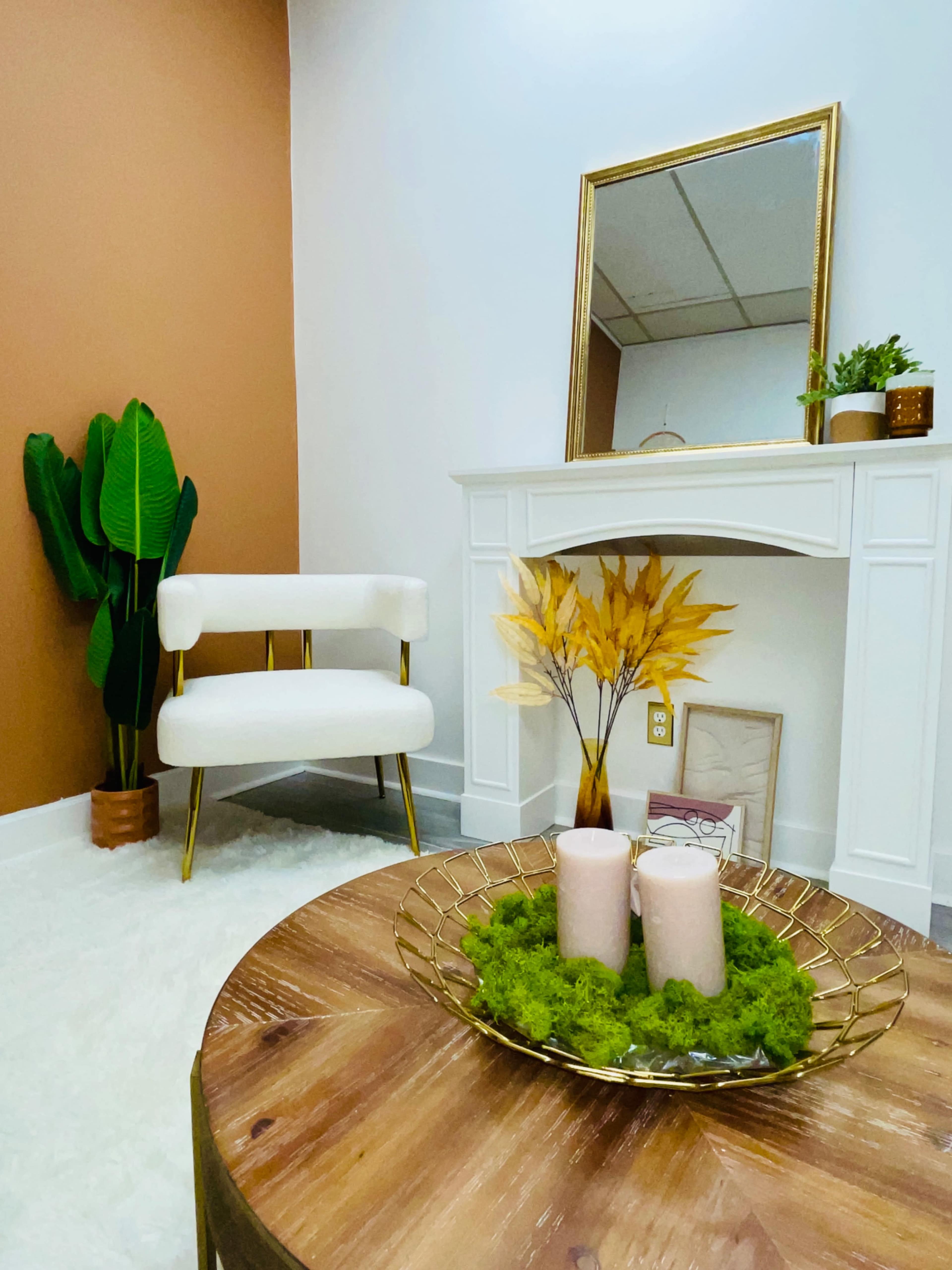 A decorated interior space featuring a white armchair, a round wooden coffee table with candles and moss, a gold-framed mirror, and a potted plant against a two-tone wall.