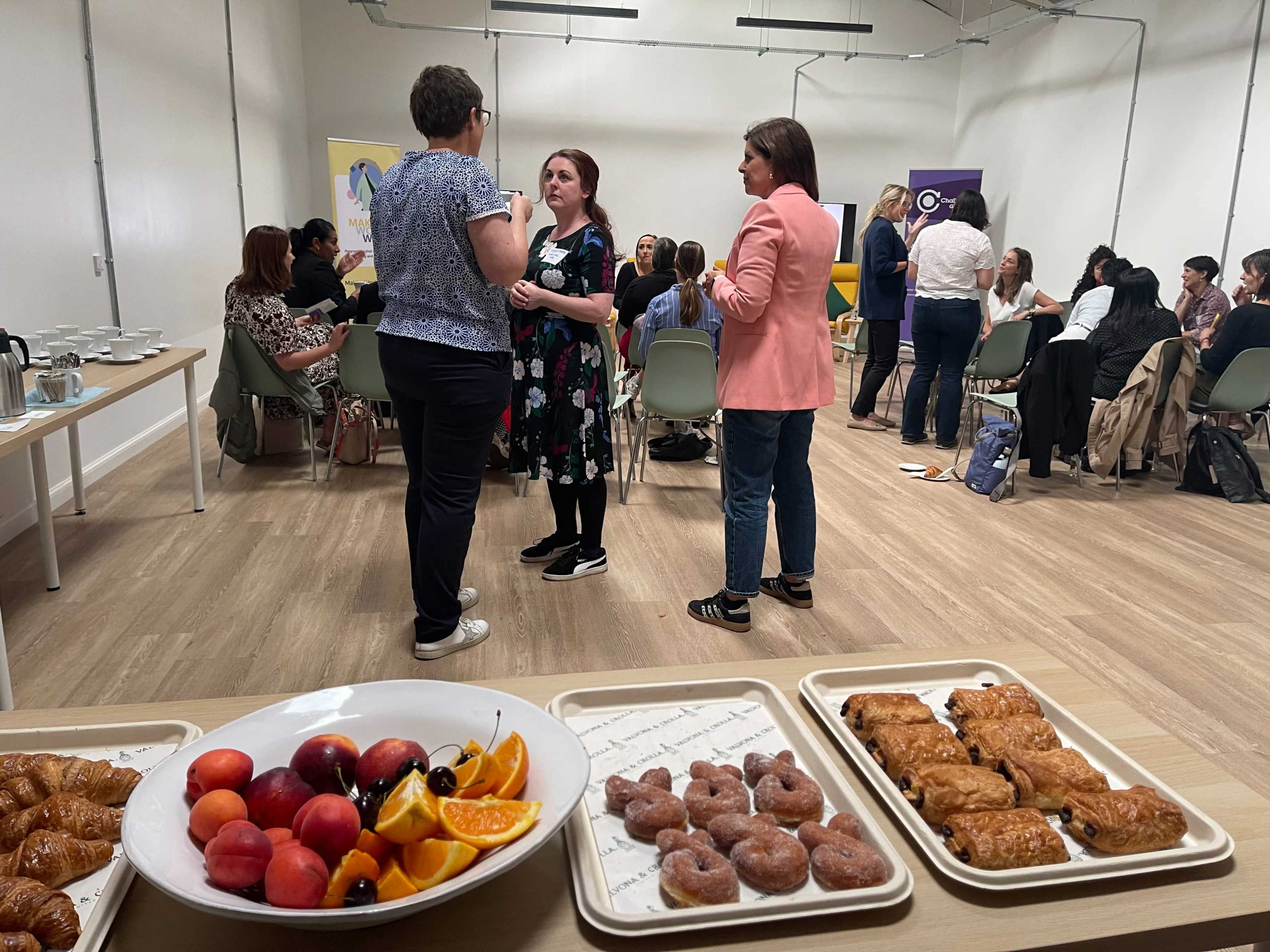 A group of people is engaged in conversation in a conference room while food items, including pastries and fruits, are displayed on a table in the foreground.